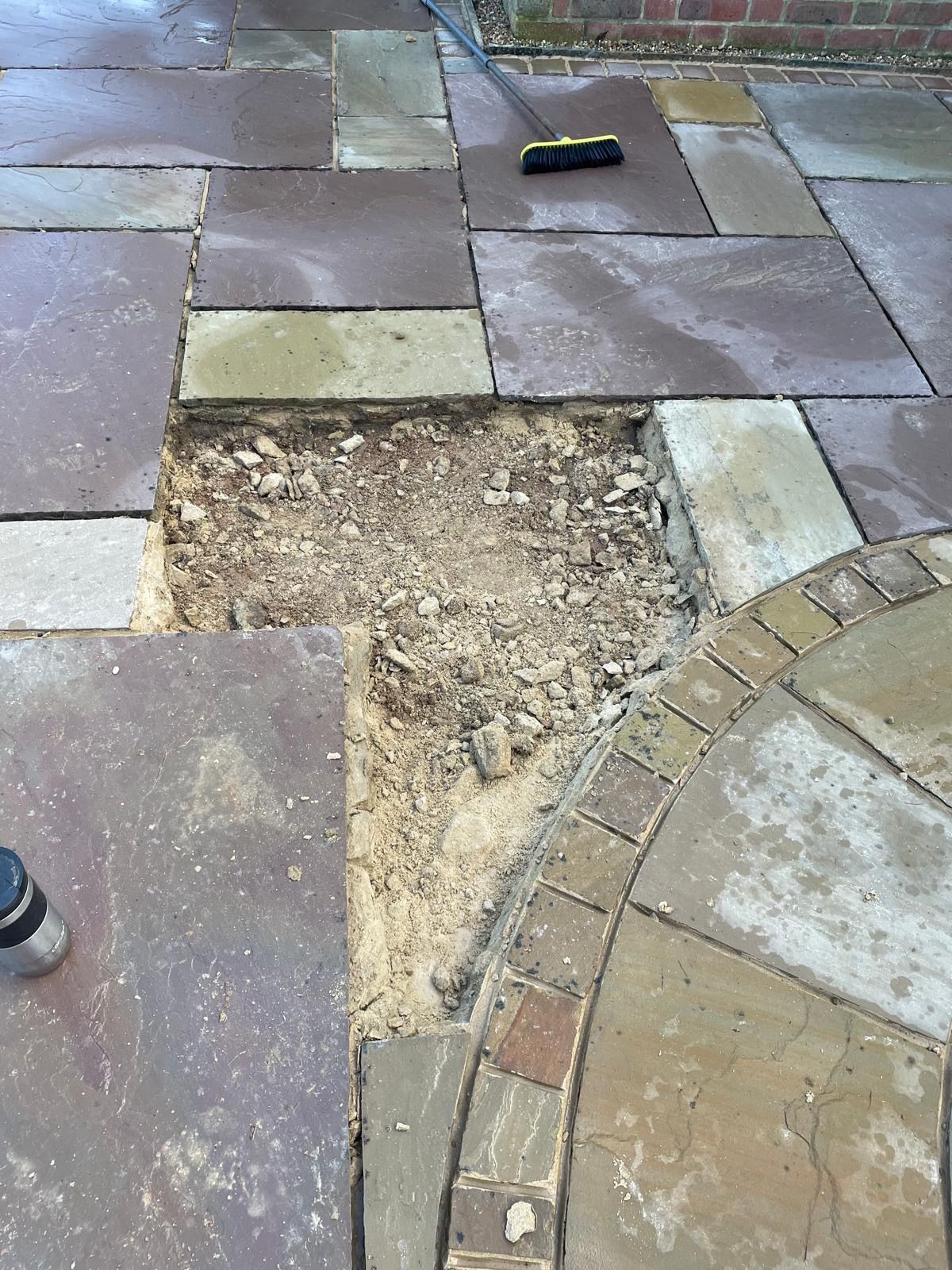 A patch of stone patio pavers is missing, revealing a light-colored sandy base and dirt in a backyard setting.