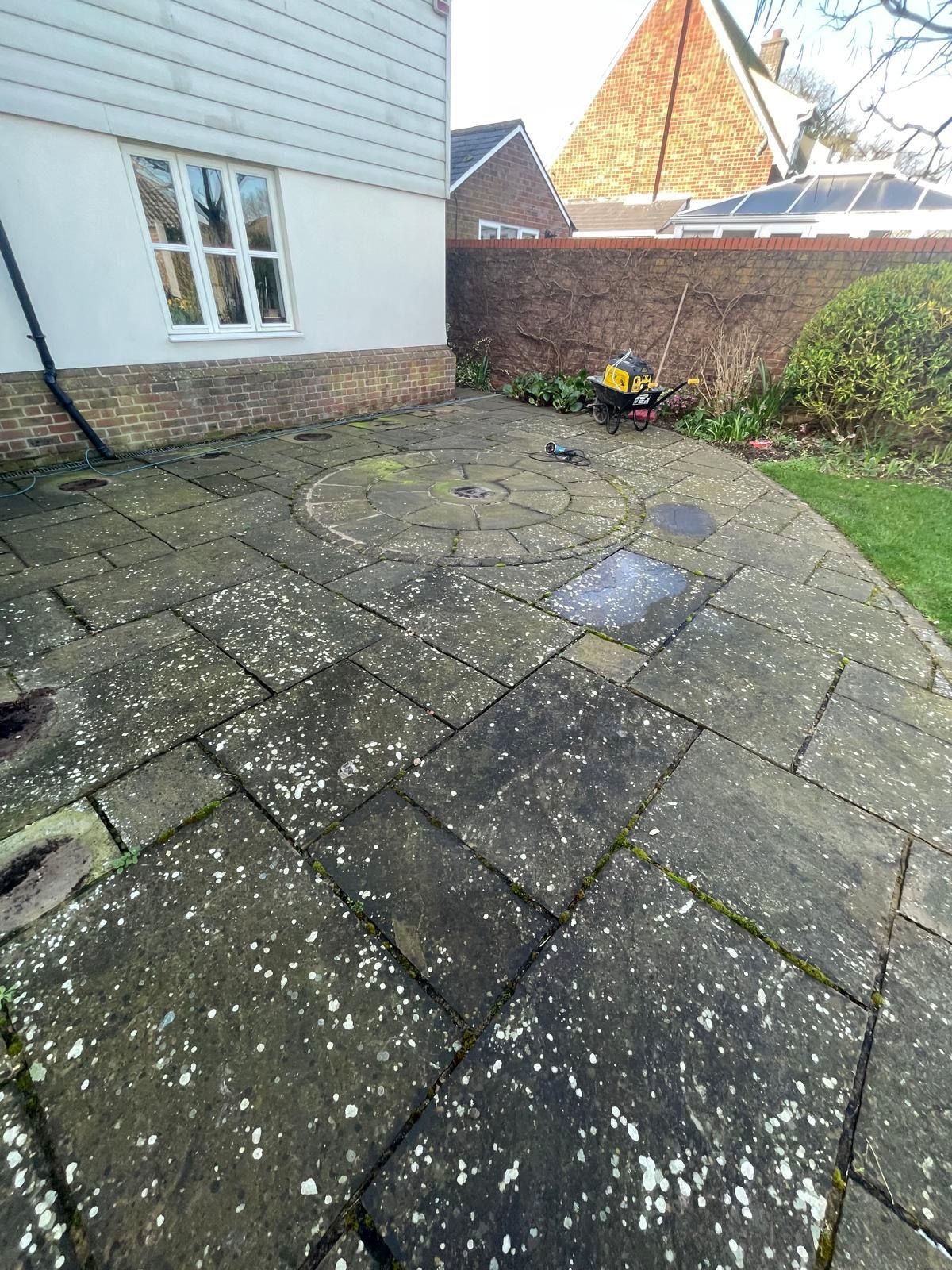 A weathered stone patio with a circular inlay, situated outside a white house with a dark brick wall in the background.