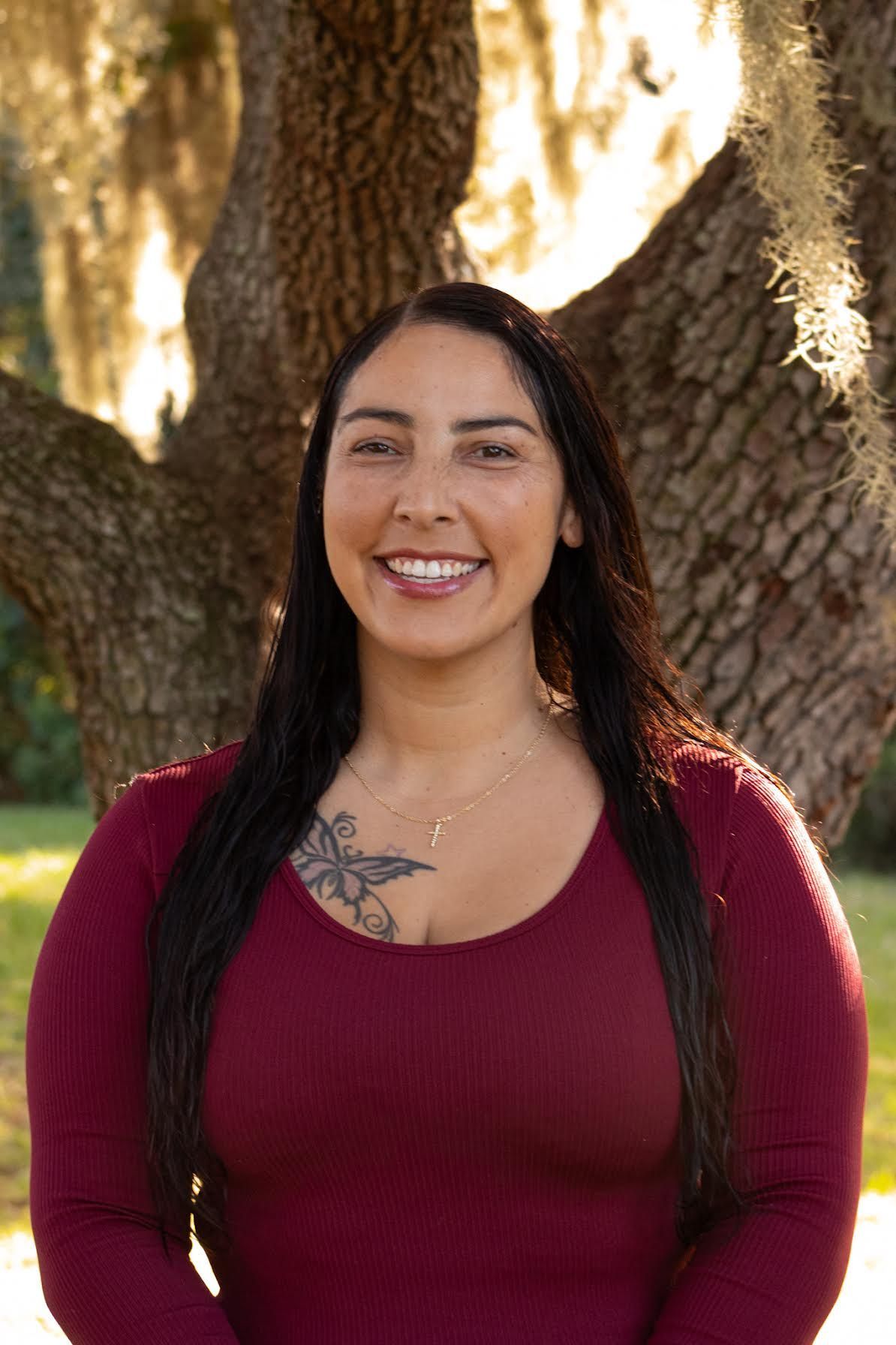 Smiling woman in a burgundy top standing outdoors in front of a large tree