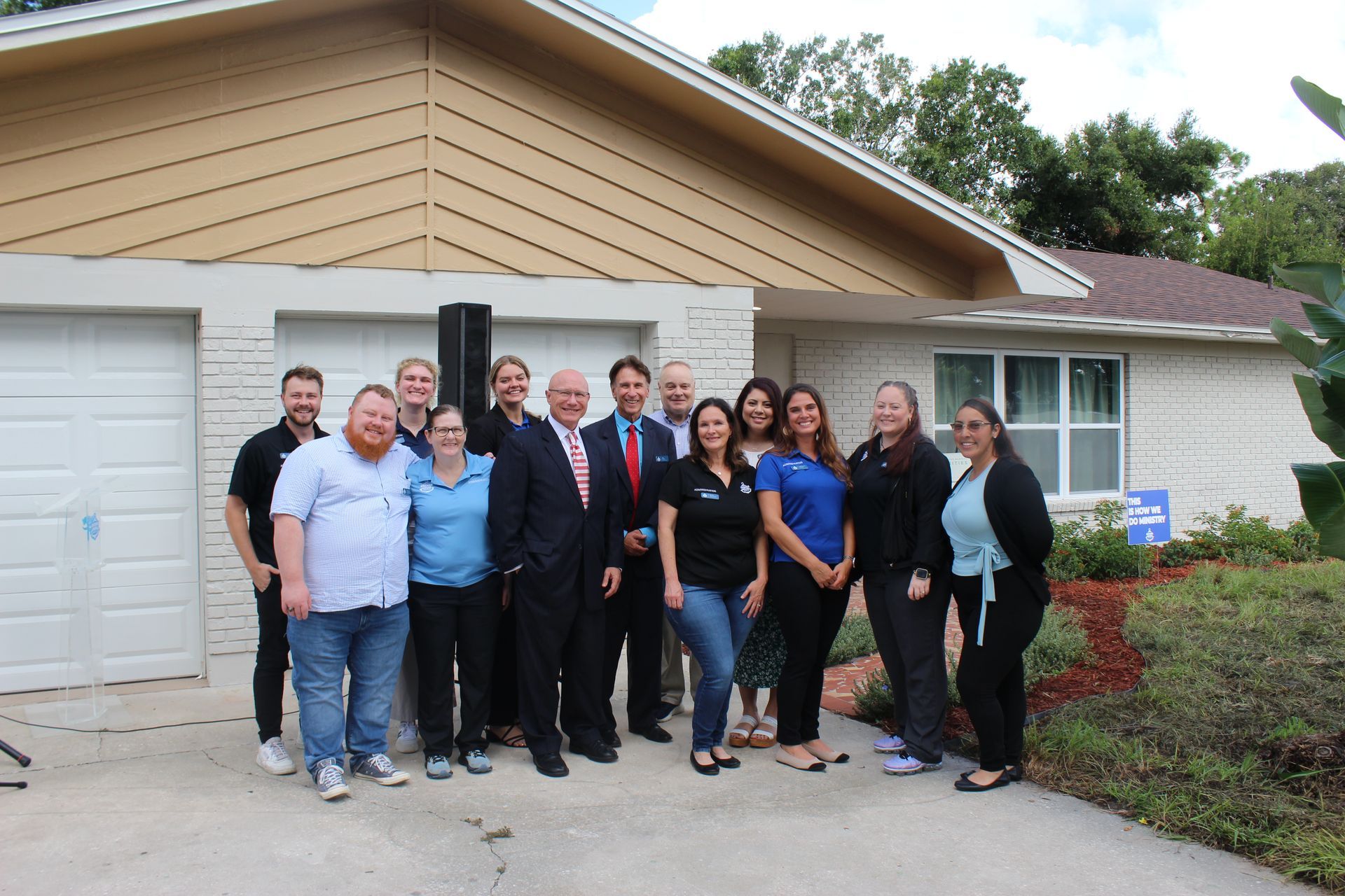 Group of people posing outside a suburban house and garage, standing on a driveway with shrubs nearby.