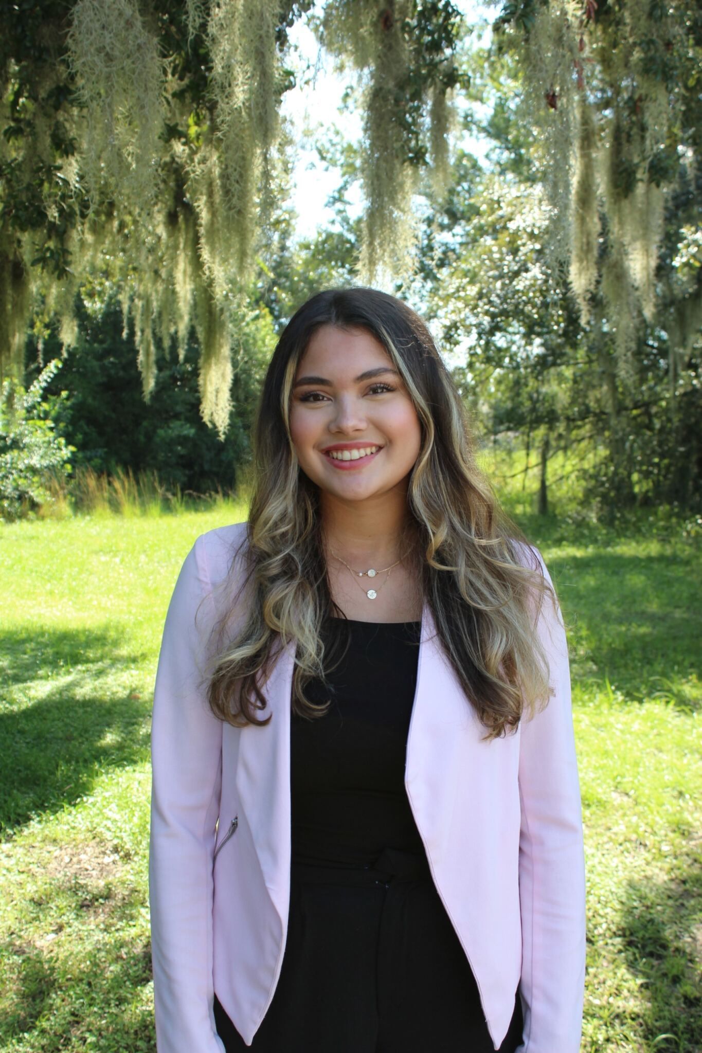 Woman smiling outdoors in a light pink blazer and black dress, standing in a sunlit park.
