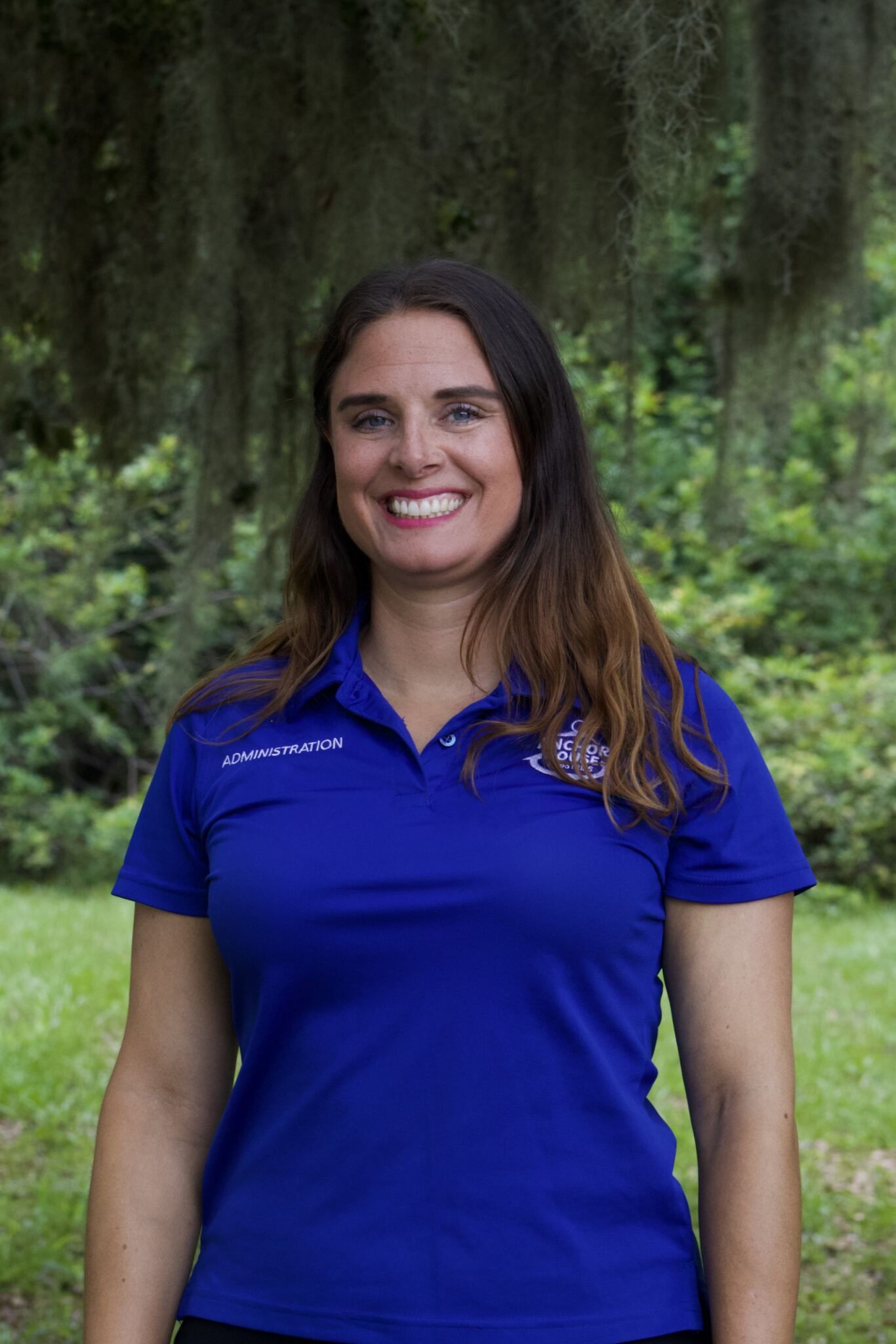 Smiling woman in a blue polo shirt standing outdoors in front of green foliage