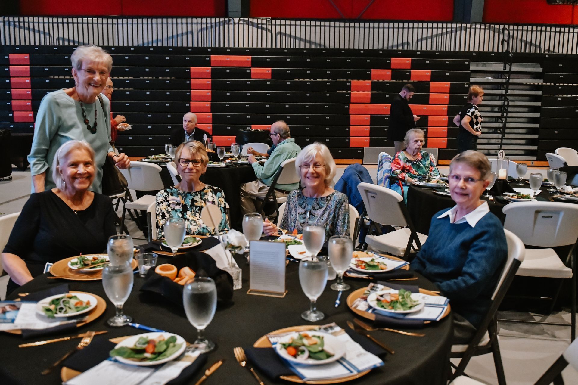 People seated at round tables in a banquet hall with red and black decor, smiling at the camera