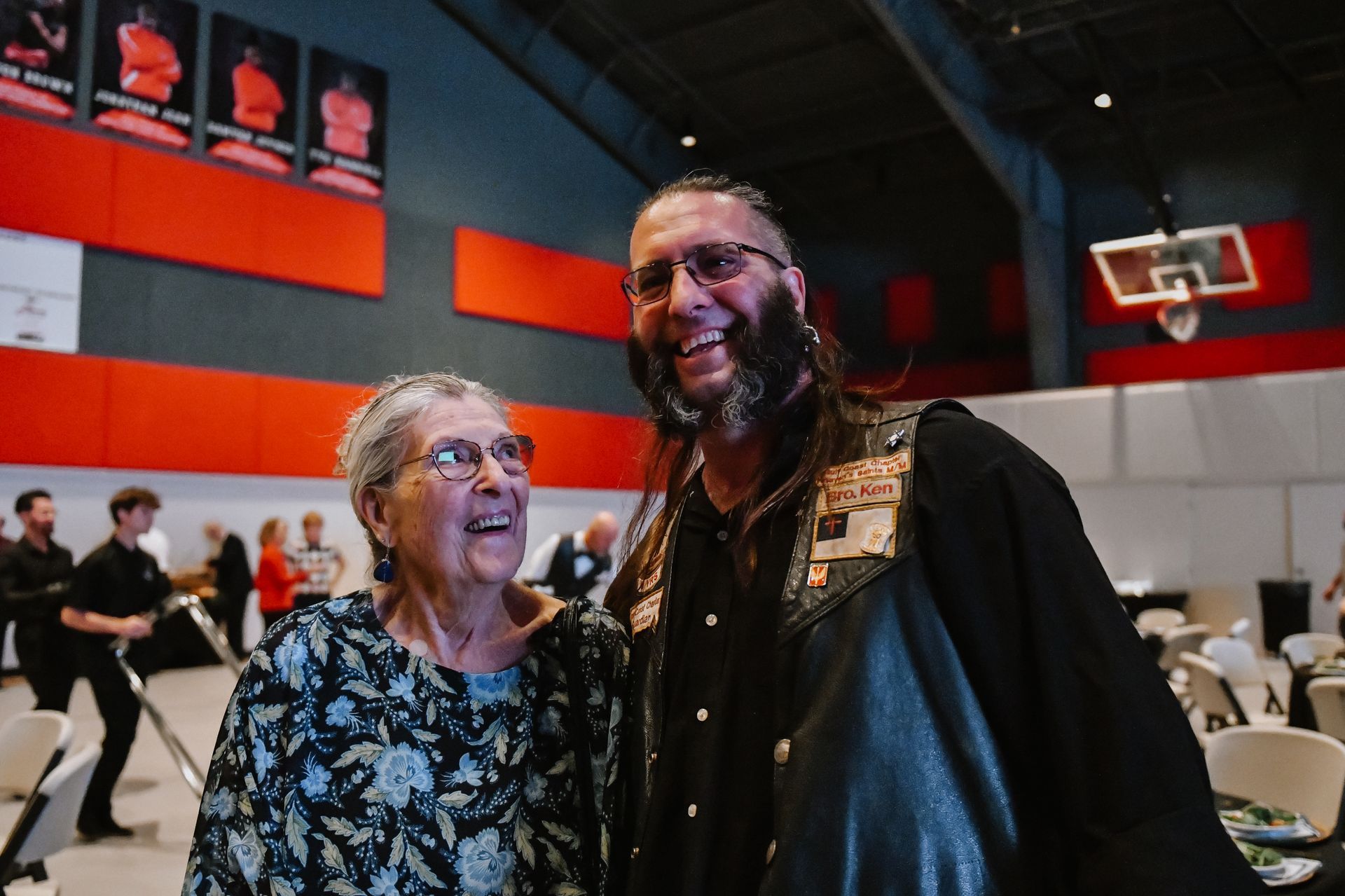 Two smiling people pose together in a gymnasium with red and orange walls.