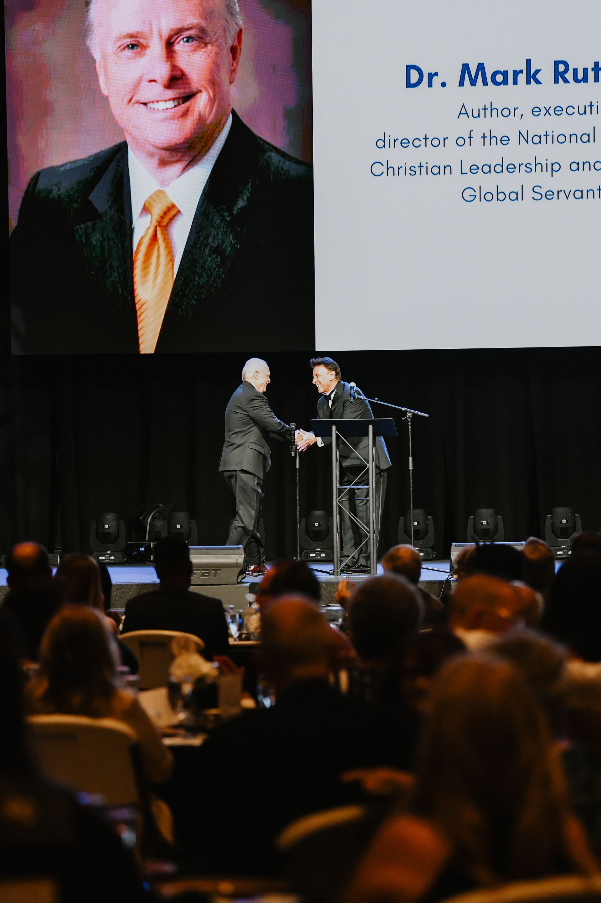 Audience watches stage presentation with speaker and large portrait on screen