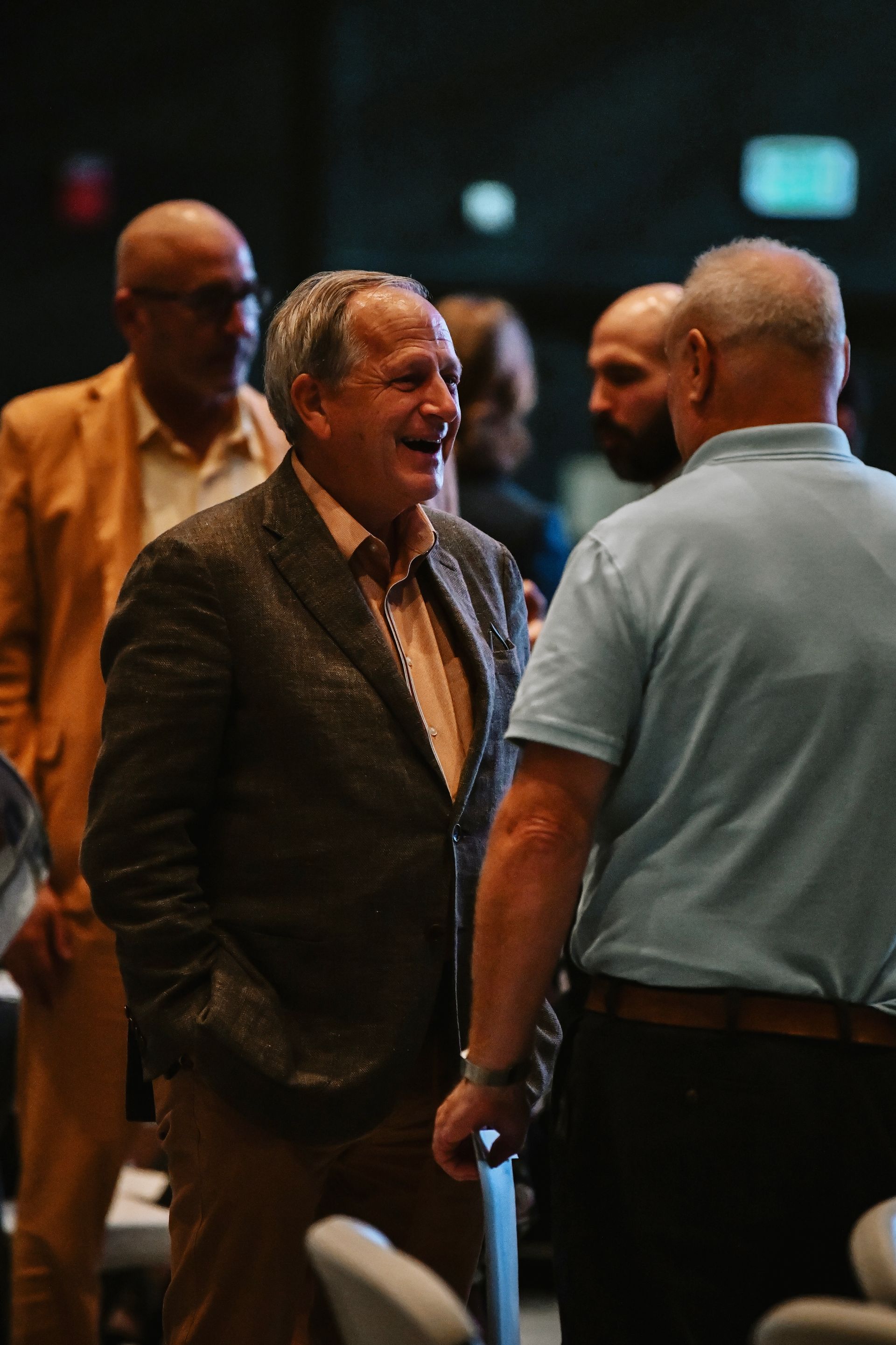 A man in a brown jacket smiles in conversation with two others at a dimly lit indoor event.