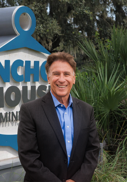 Smiling man in a dark suit and blue shirt standing beside a sign outdoors