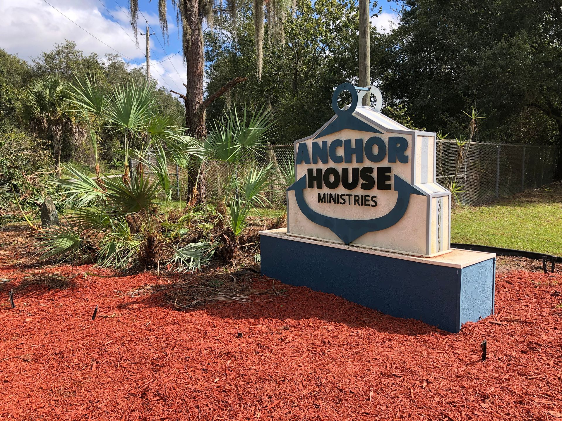 Anchor House Ministries sign beside red mulch and tropical plants outdoors