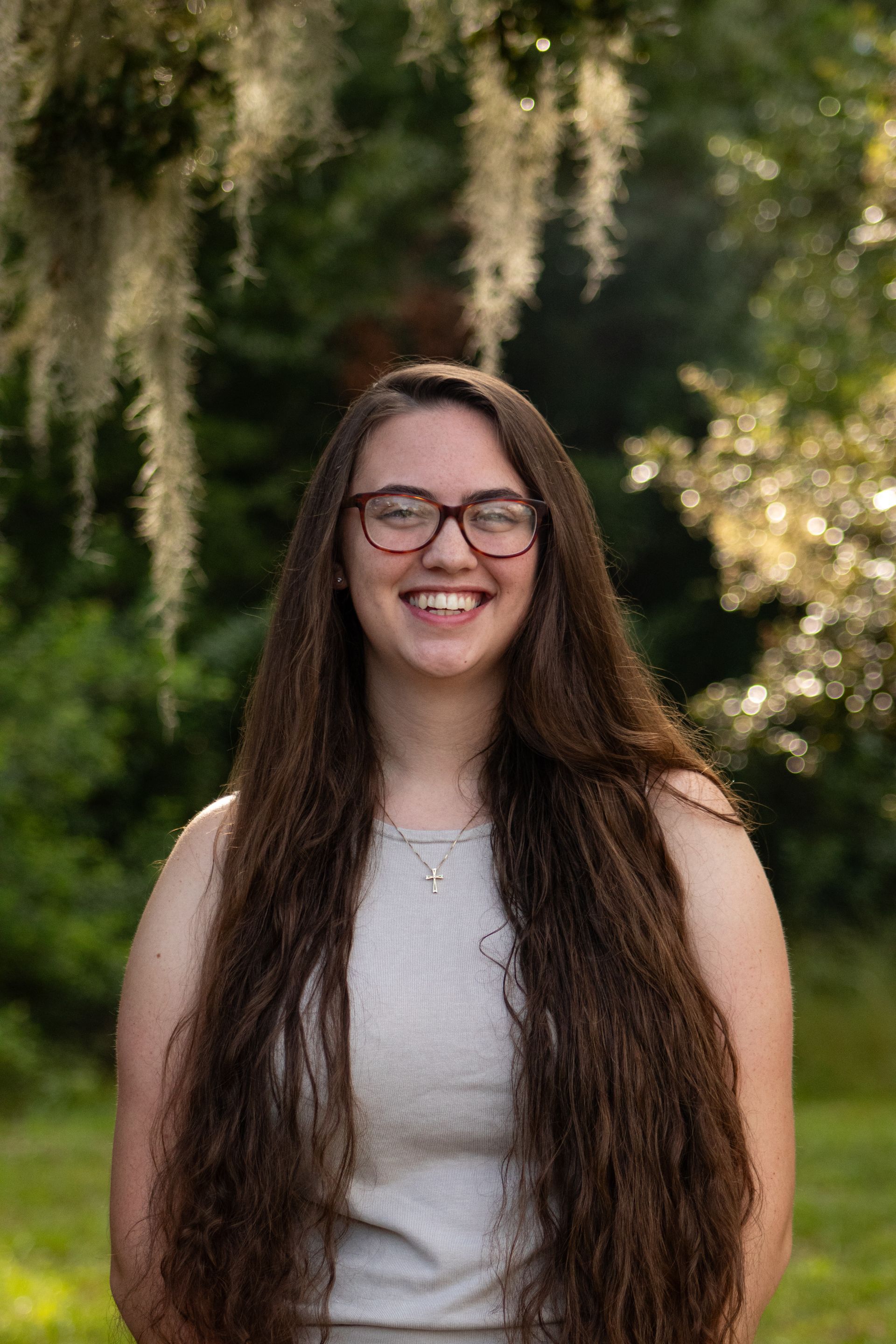 Smiling person with long dark hair and glasses standing outdoors before a blurred green background