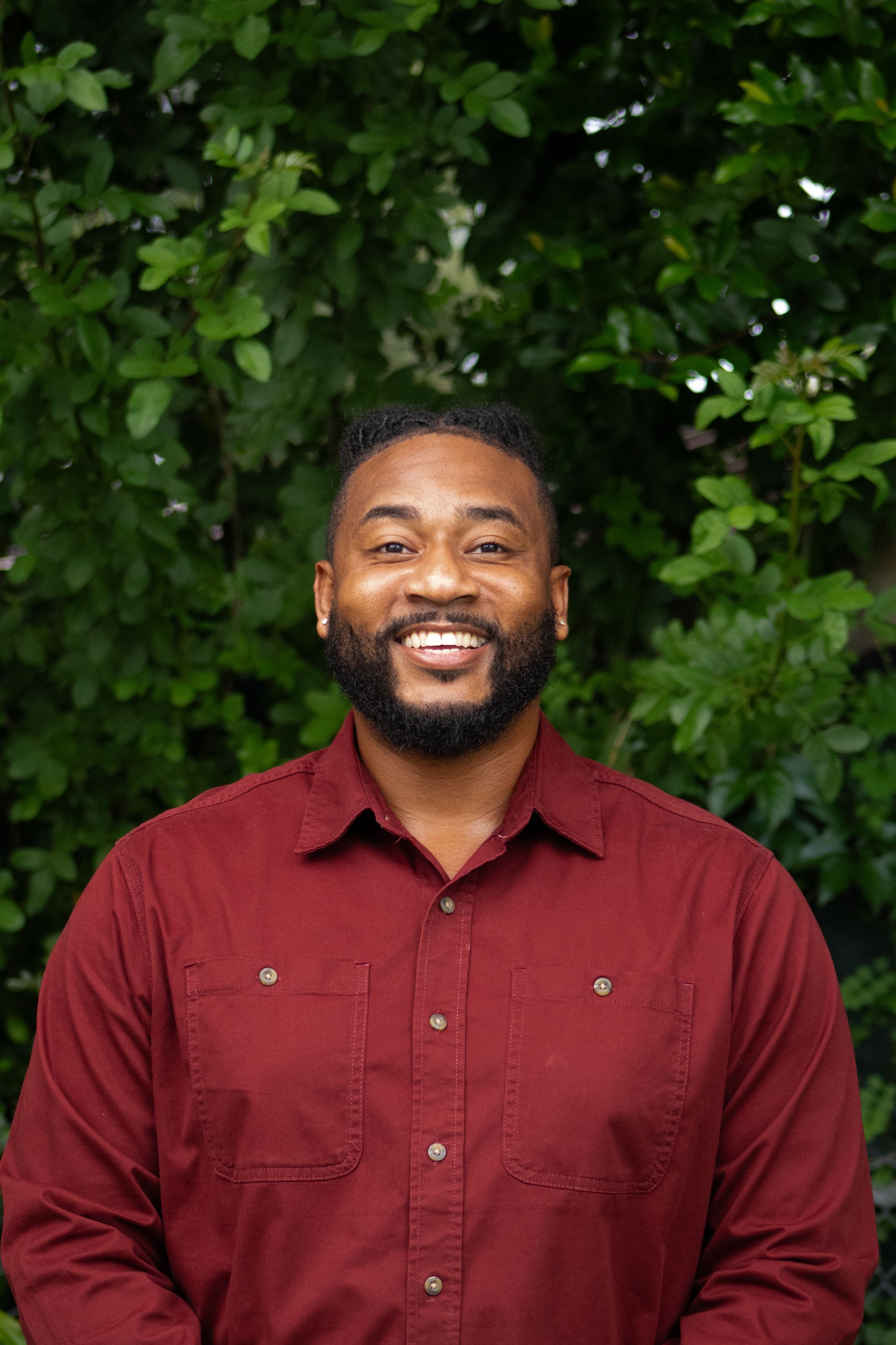 Smiling man in a maroon shirt standing before green foliage