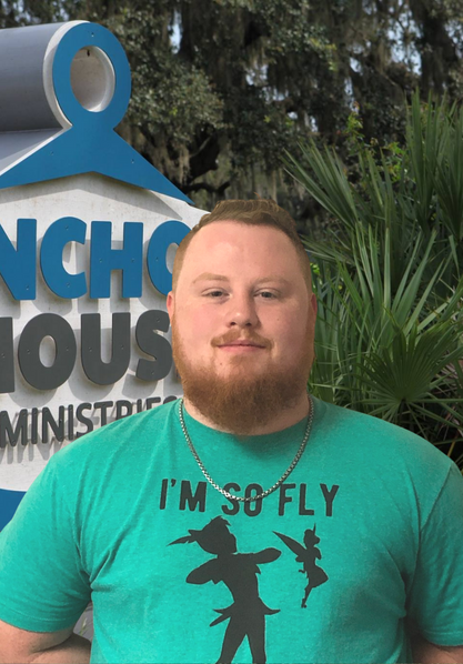 Man in teal T-shirt standing in front of an outdoor ministry sign and greenery