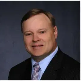 Studio headshot of a man in a dark suit, light blue shirt, and patterned tie against a dark background