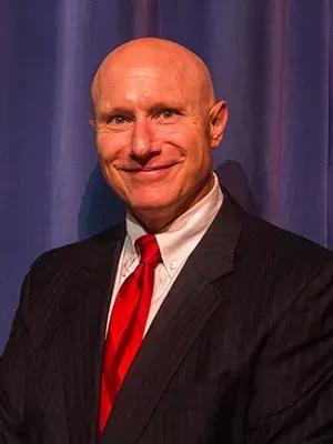 Smiling bald man in a dark suit and red tie against a blue curtain backdrop