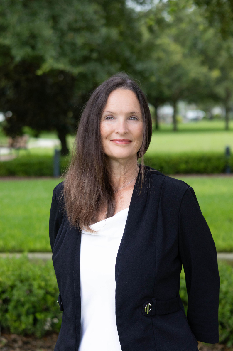 Woman standing outdoors in a park, wearing a black blazer and white shirt, with trees and grass behind her