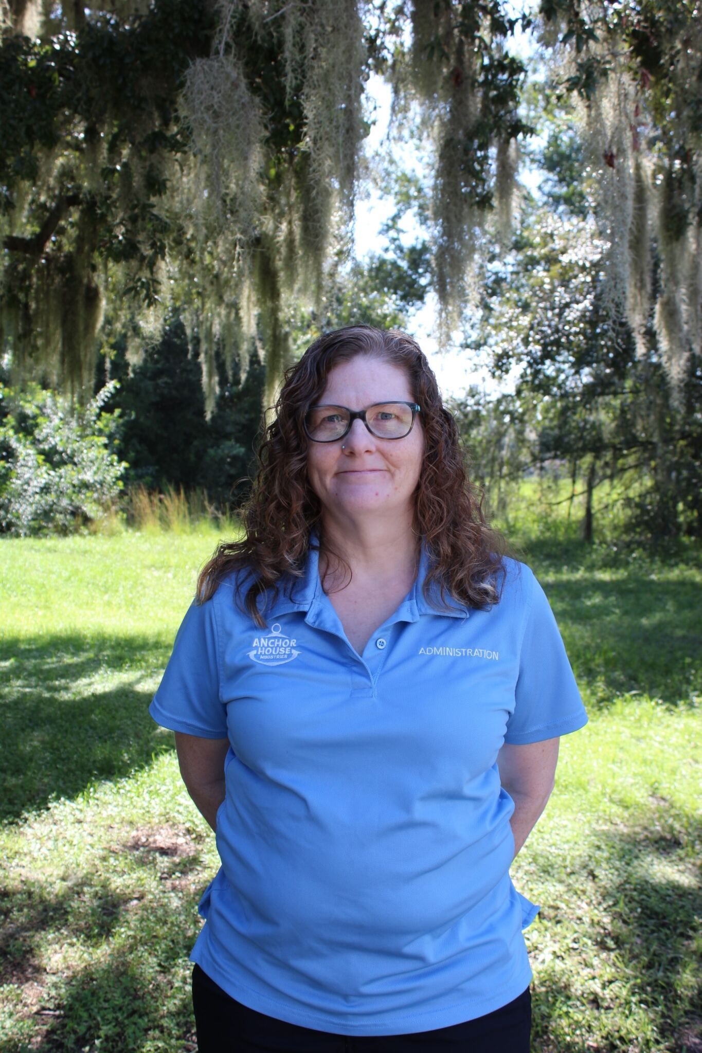 Person in a blue polo shirt standing outdoors in a sunny park with hanging Spanish moss.