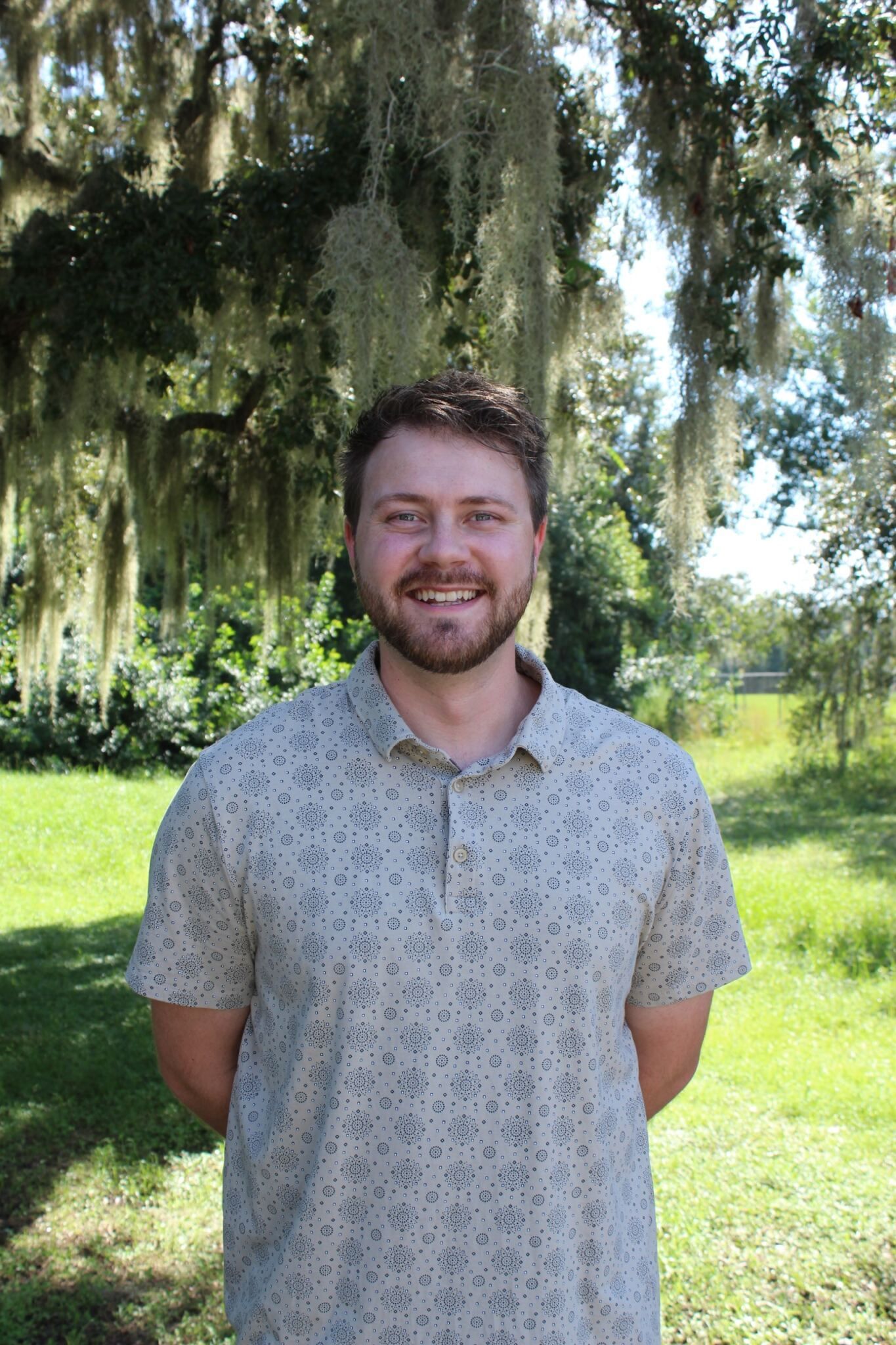 Smiling person standing outdoors in a sunny park with trees and grass in the background.