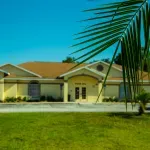 Single-story yellow house with a tiled roof, palm fronds in front, and a green lawn under a blue sky