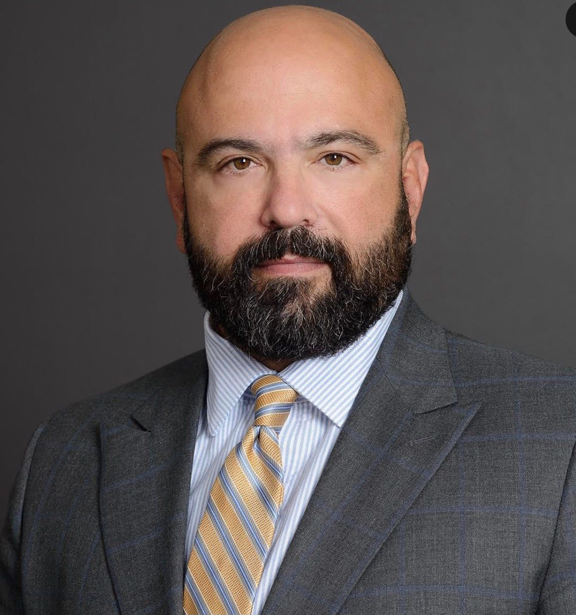 Studio portrait of a bald man with a beard in a gray suit and striped tie on a dark background