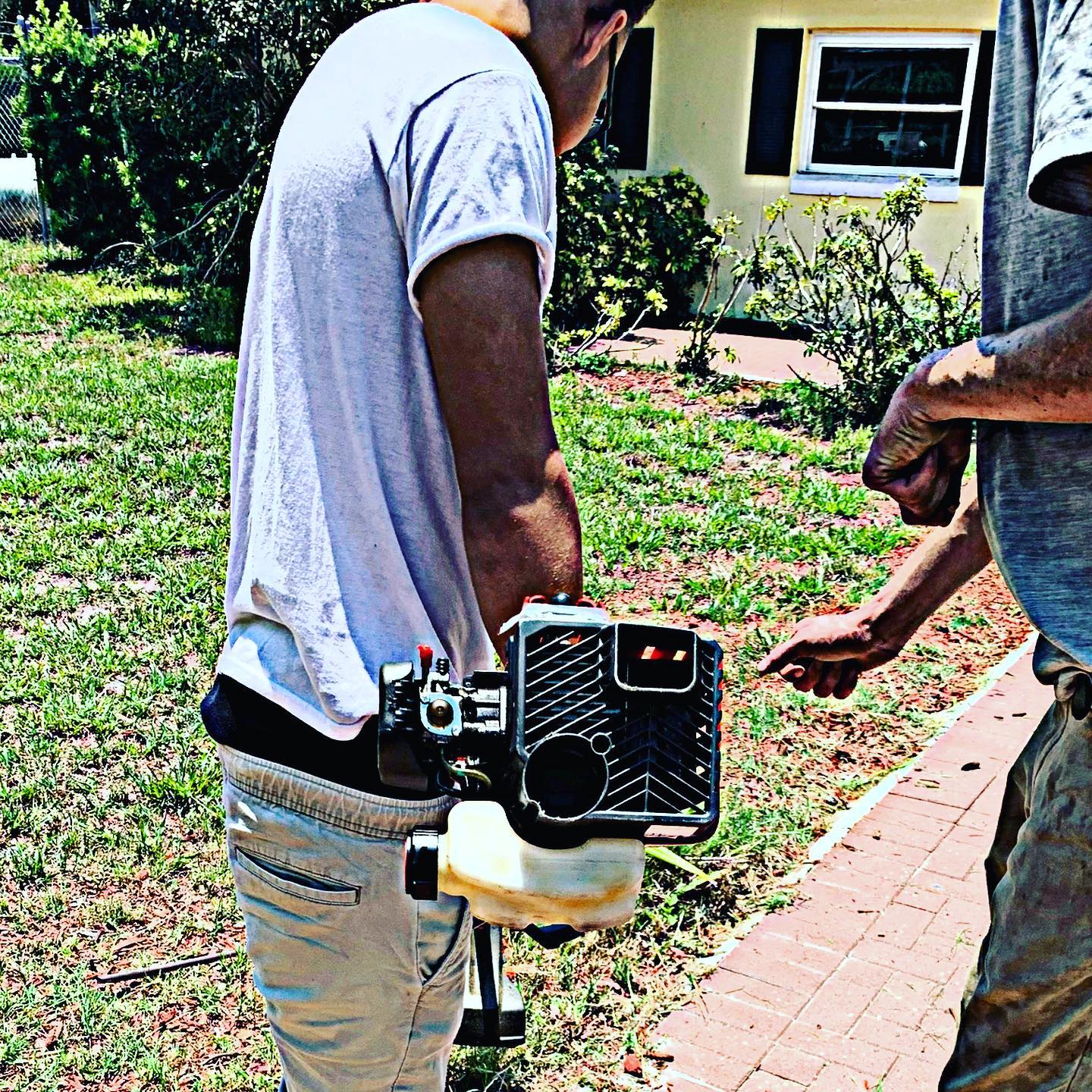 Two people handle a lawn edger on a brick path beside a house and garden.