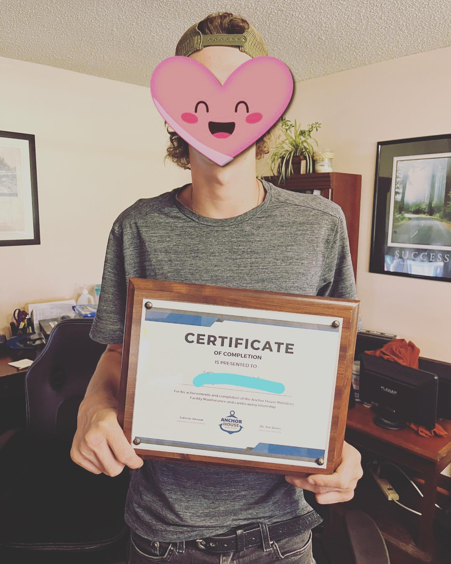 Person holding a framed certificate indoors, wearing a gray shirt and smiling heart-face emoji mask