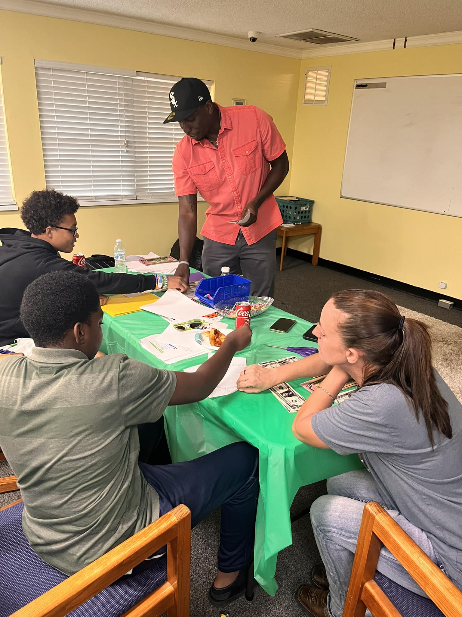 People seated around a table in a classroom, working with supplies while one person stands nearby.