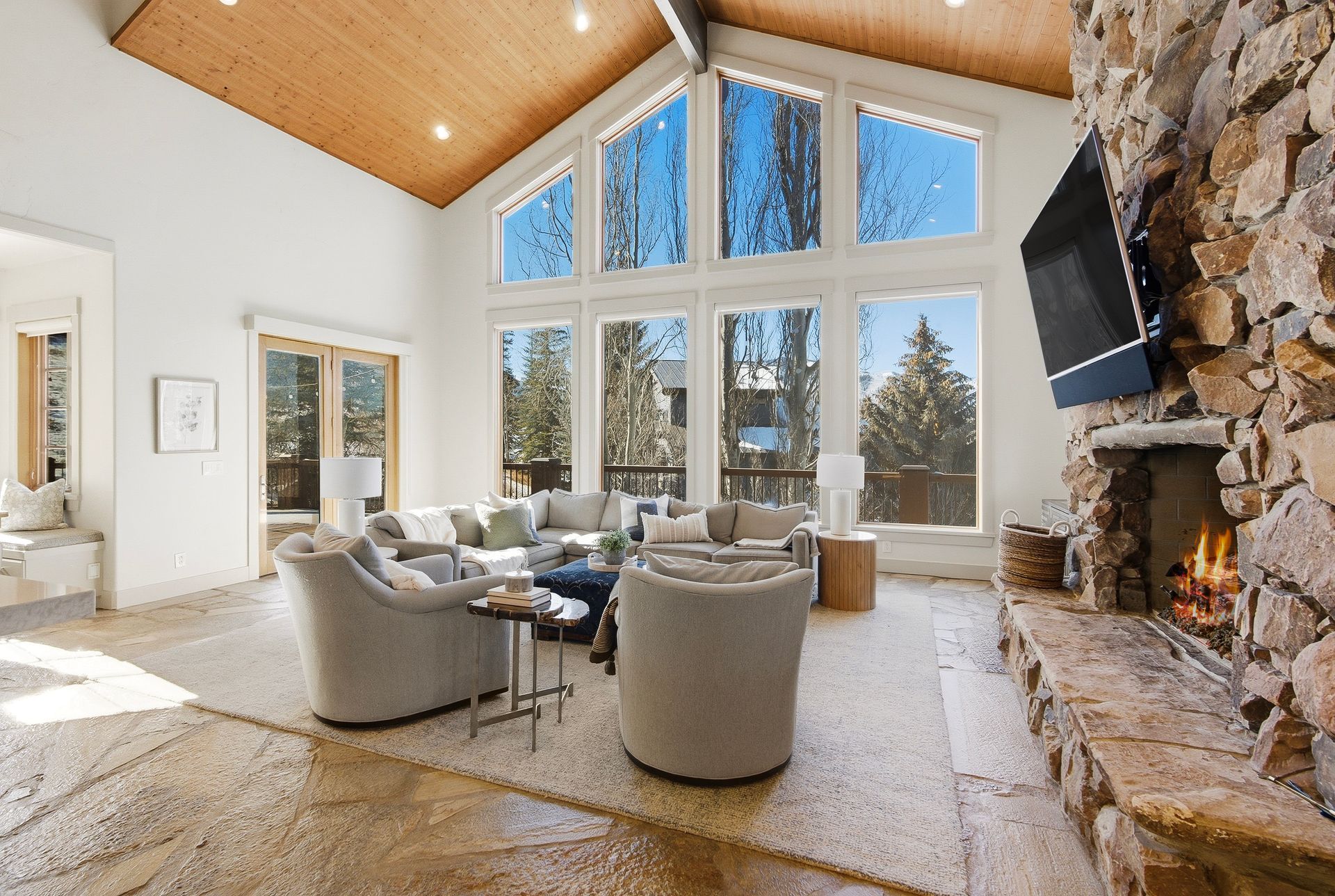 Spacious living room with vaulted wood ceiling, stone fireplace, large windows, and gray seating.