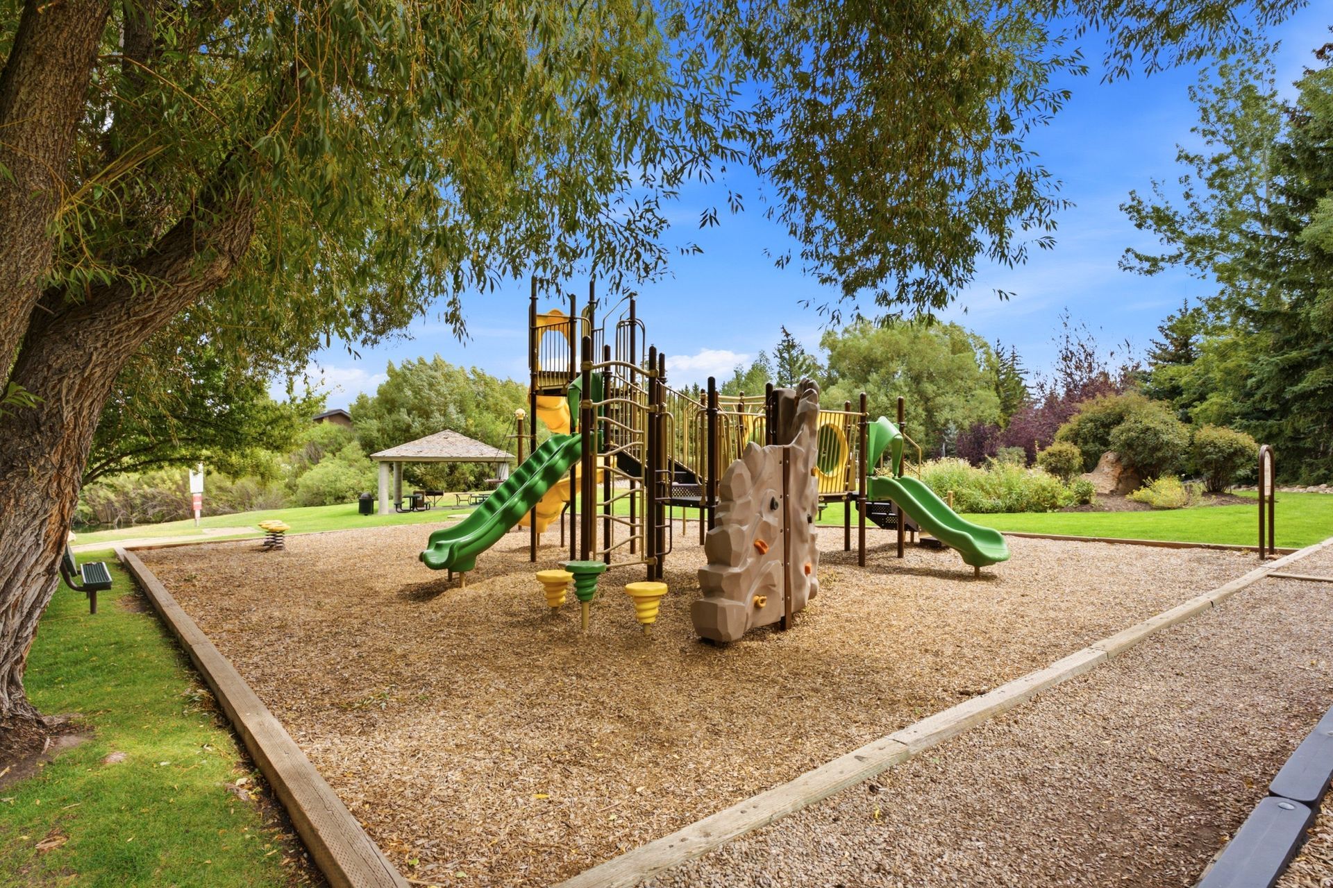 Playground with slides and climbing structure in a park, bordered by trees and grass.