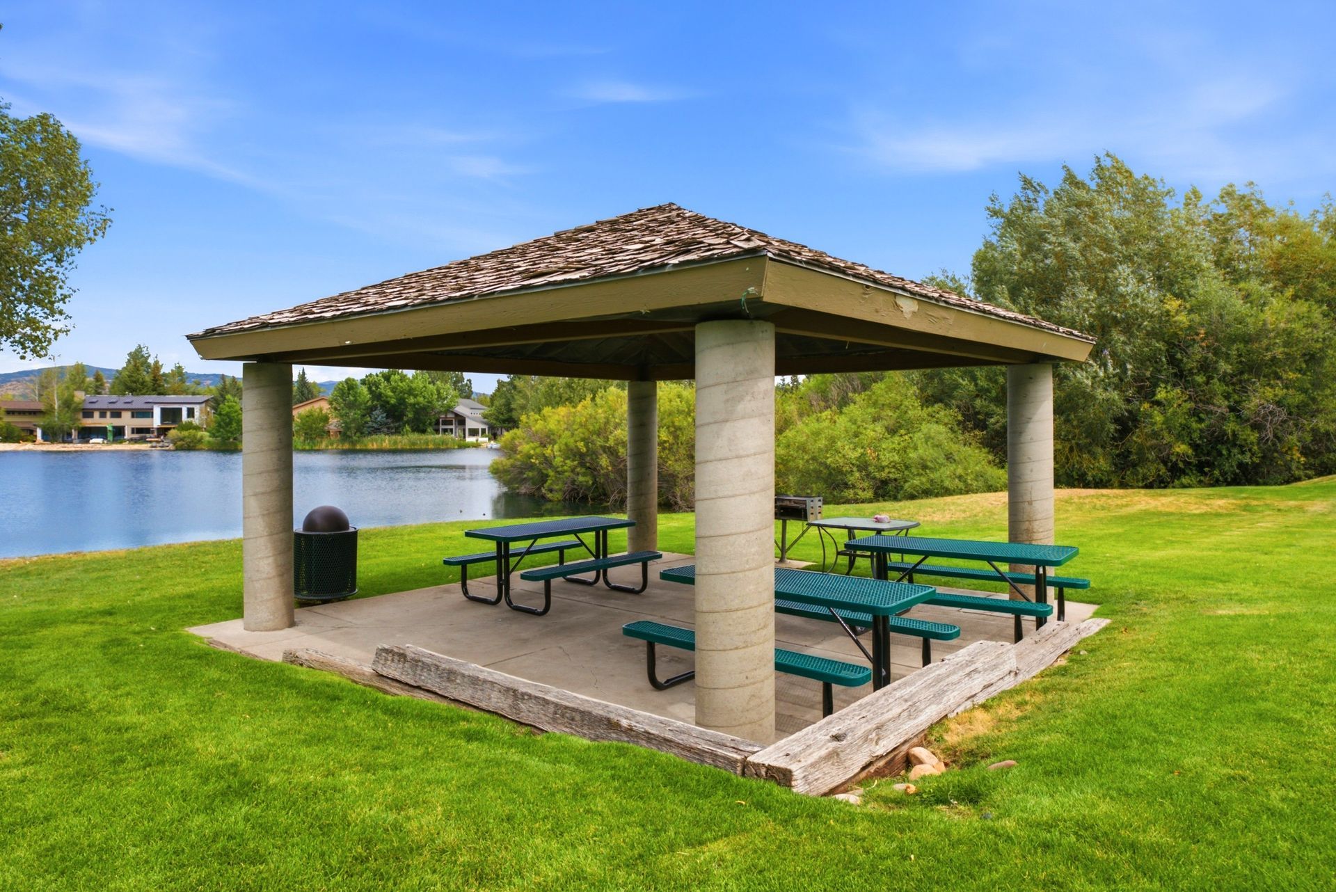 Open-air picnic shelter with green tables beside a lake, surrounded by grass and trees
