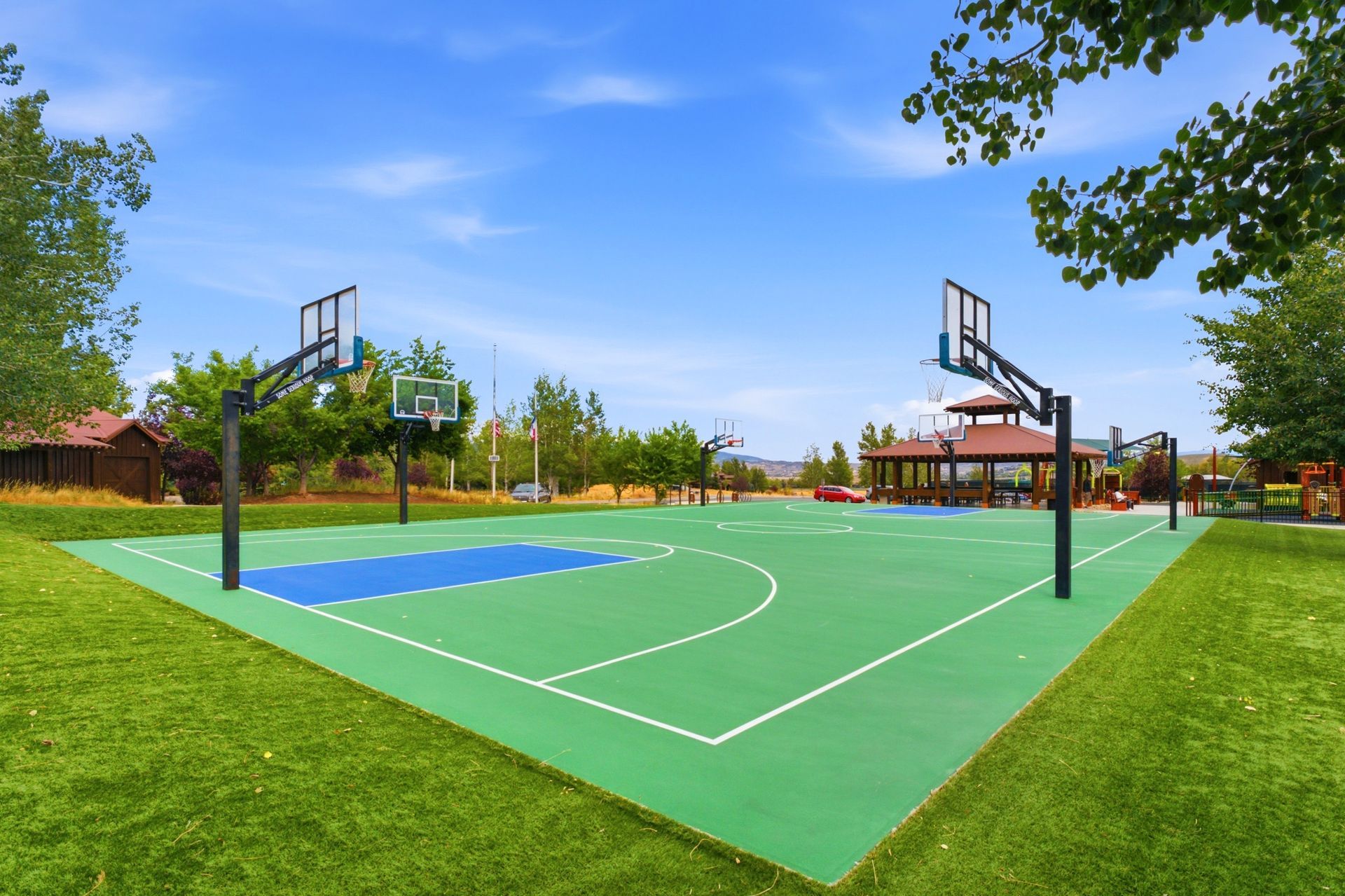 Outdoor basketball court with green and blue flooring, hoops, and trees under a clear blue sky