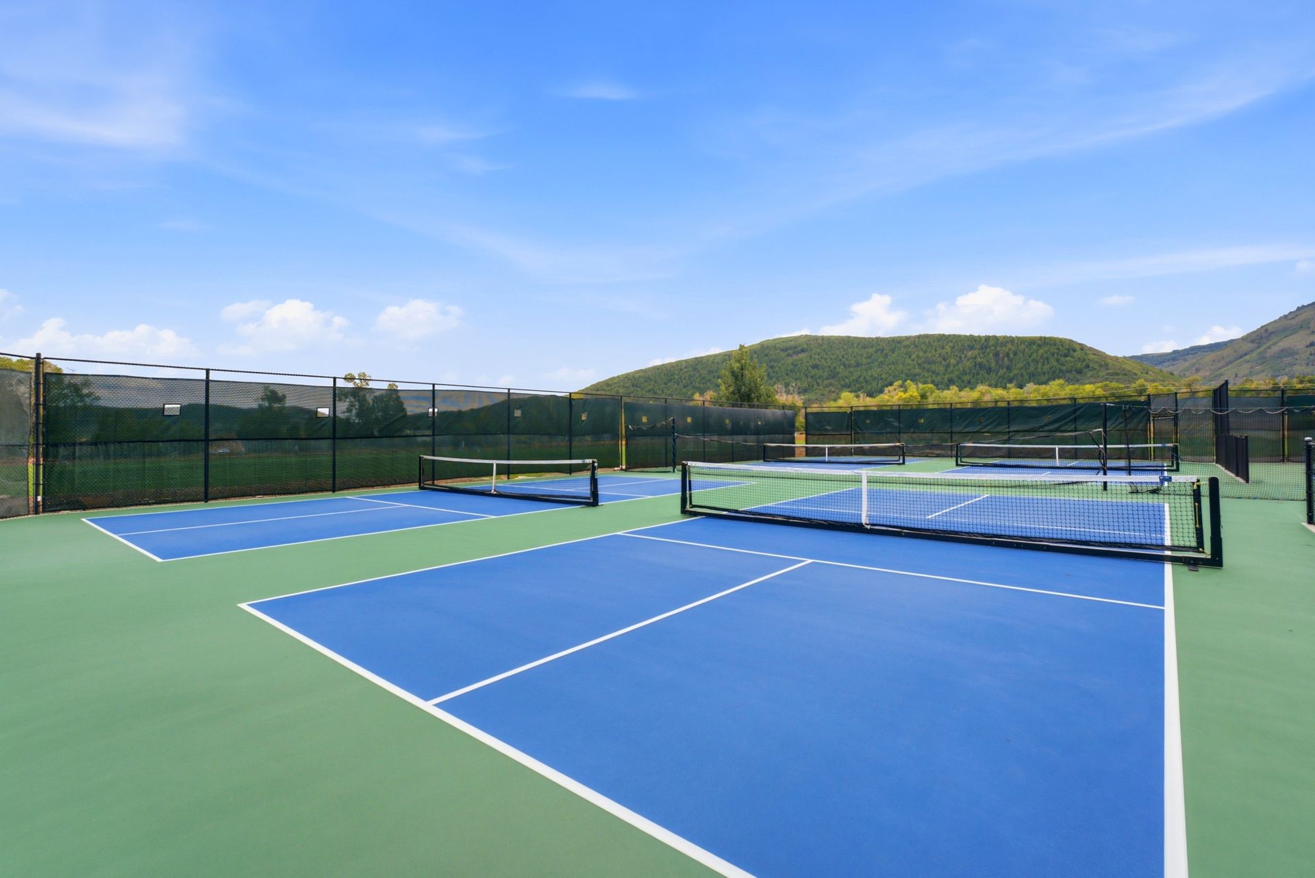Blue and green outdoor tennis courts with nets, hills, and a clear sky in the background