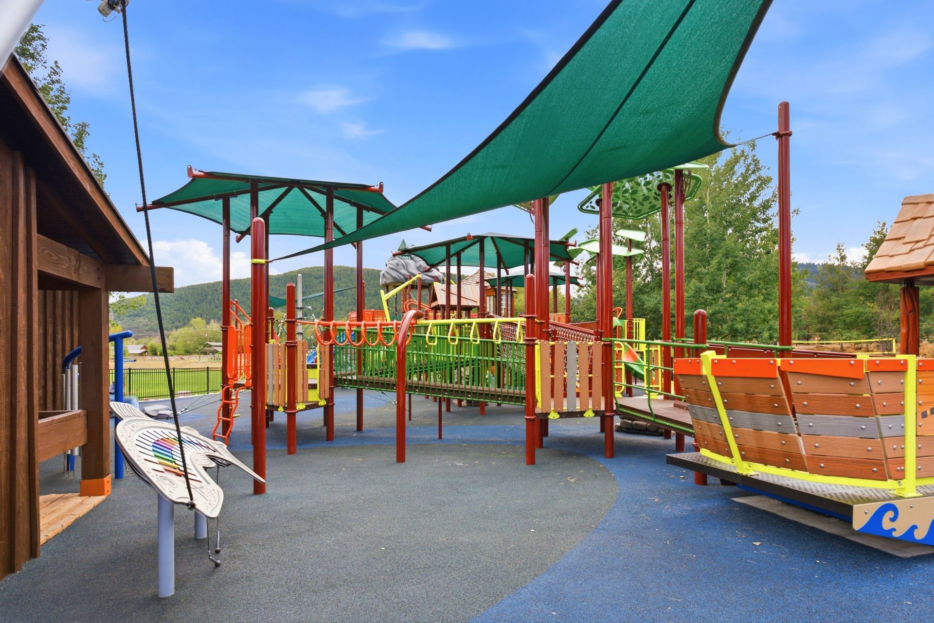 Outdoor playground with green shade sails, climbing structures, and rubber flooring under a blue sky