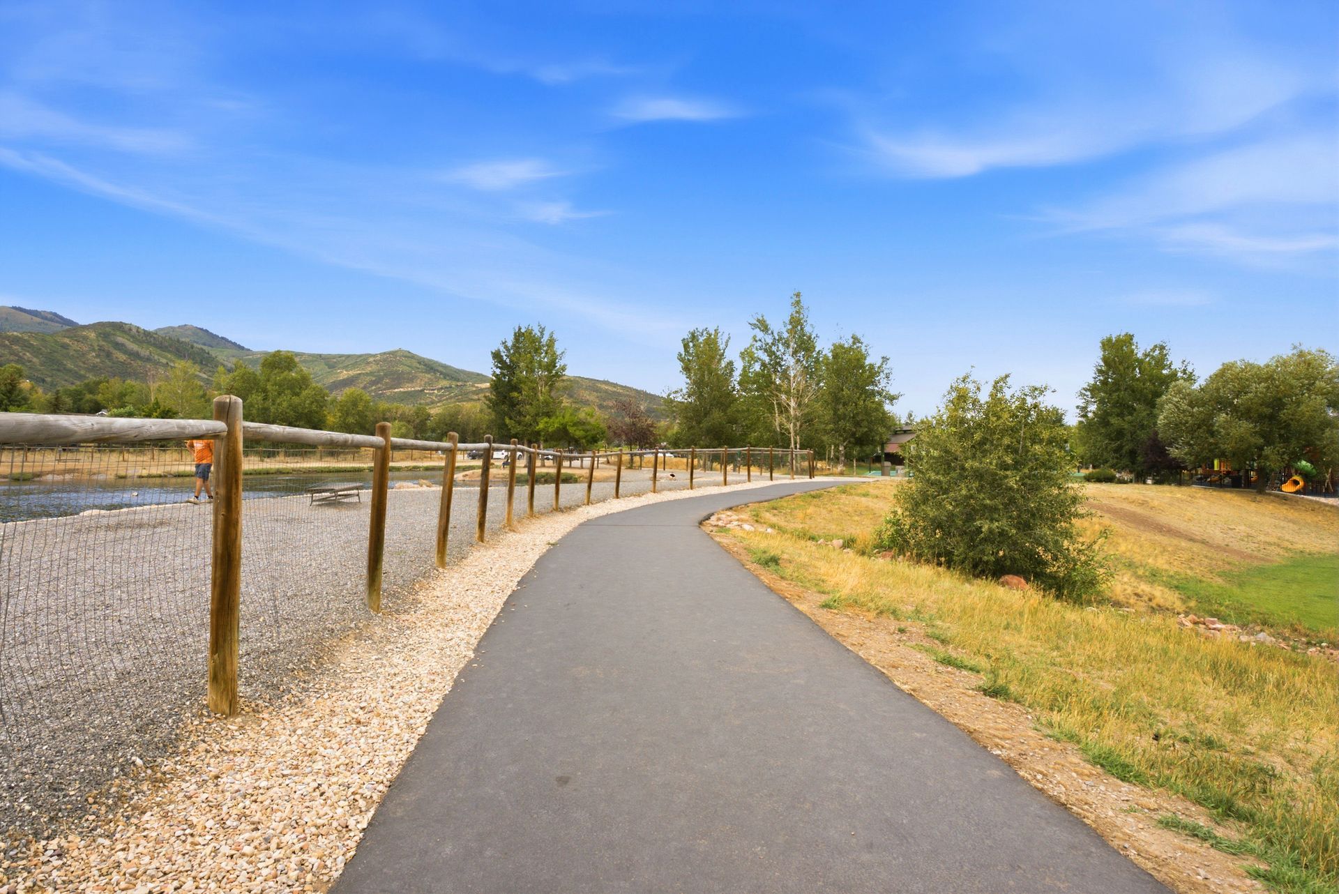 Paved path winding through a grassy park with wooden fence, trees, and mountains under a blue sky