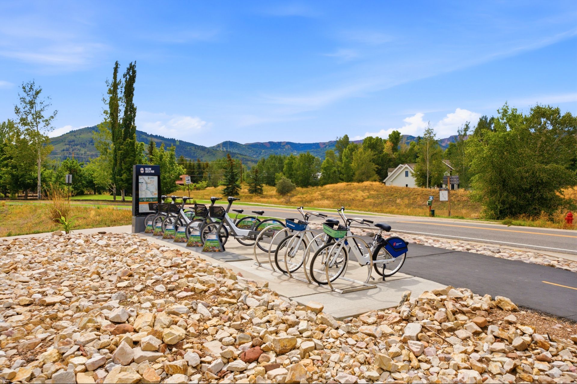 Bike-share station with several parked bicycles beside a paved path and rocky landscaping, with mountains in the background
