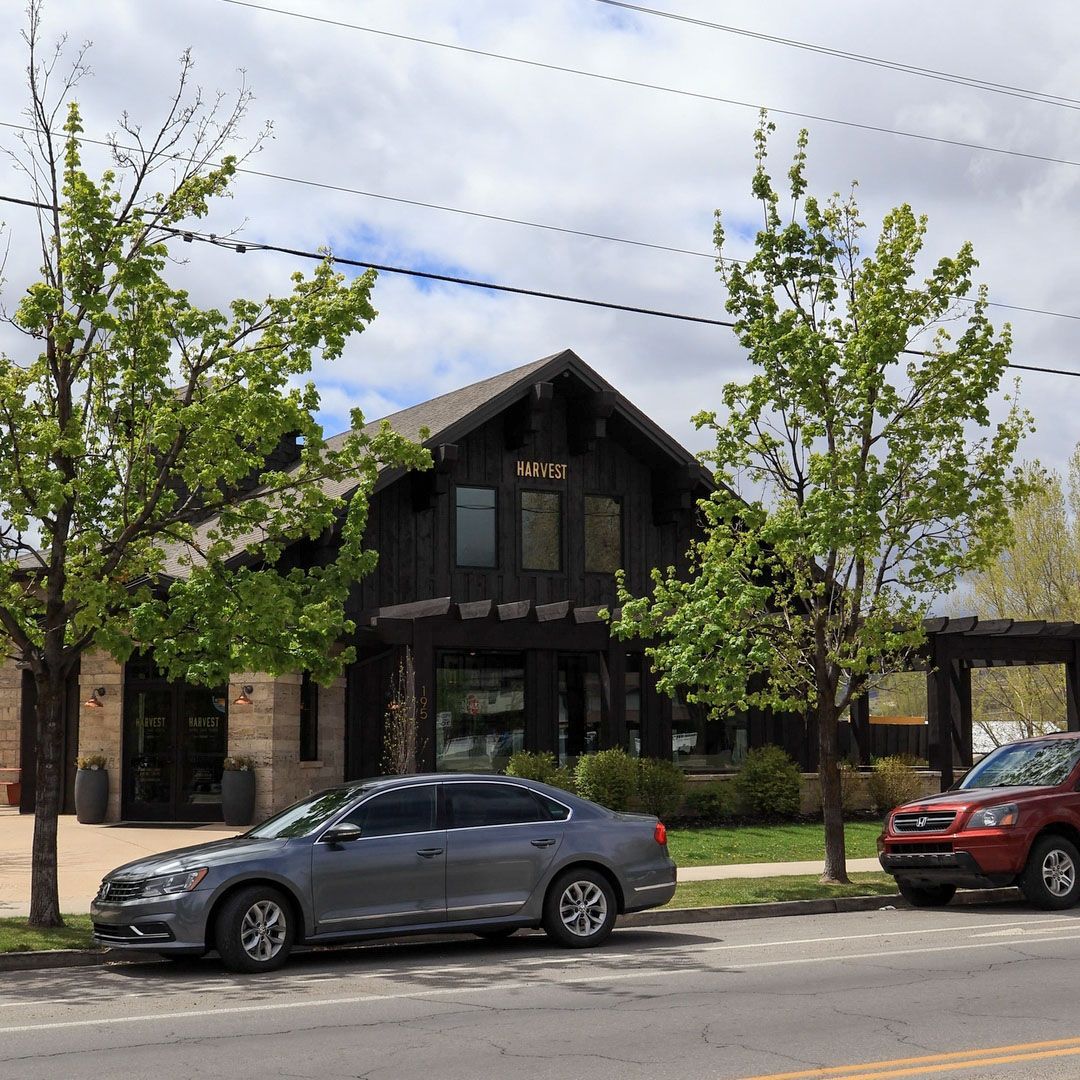 Exterior view of the Harvest cafe location in Midway, Utah, featuring a rustic dark wood and stone storefront with gold lettering, framed by green trees on a sunny day.