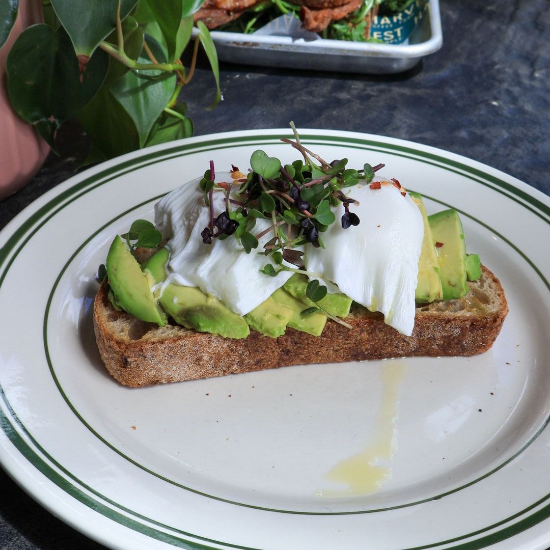 A close-up of a thick slice of sourdough avocado toast topped with a poached egg, fresh microgreens, and red pepper flakes, served on a white plate with green stripes.