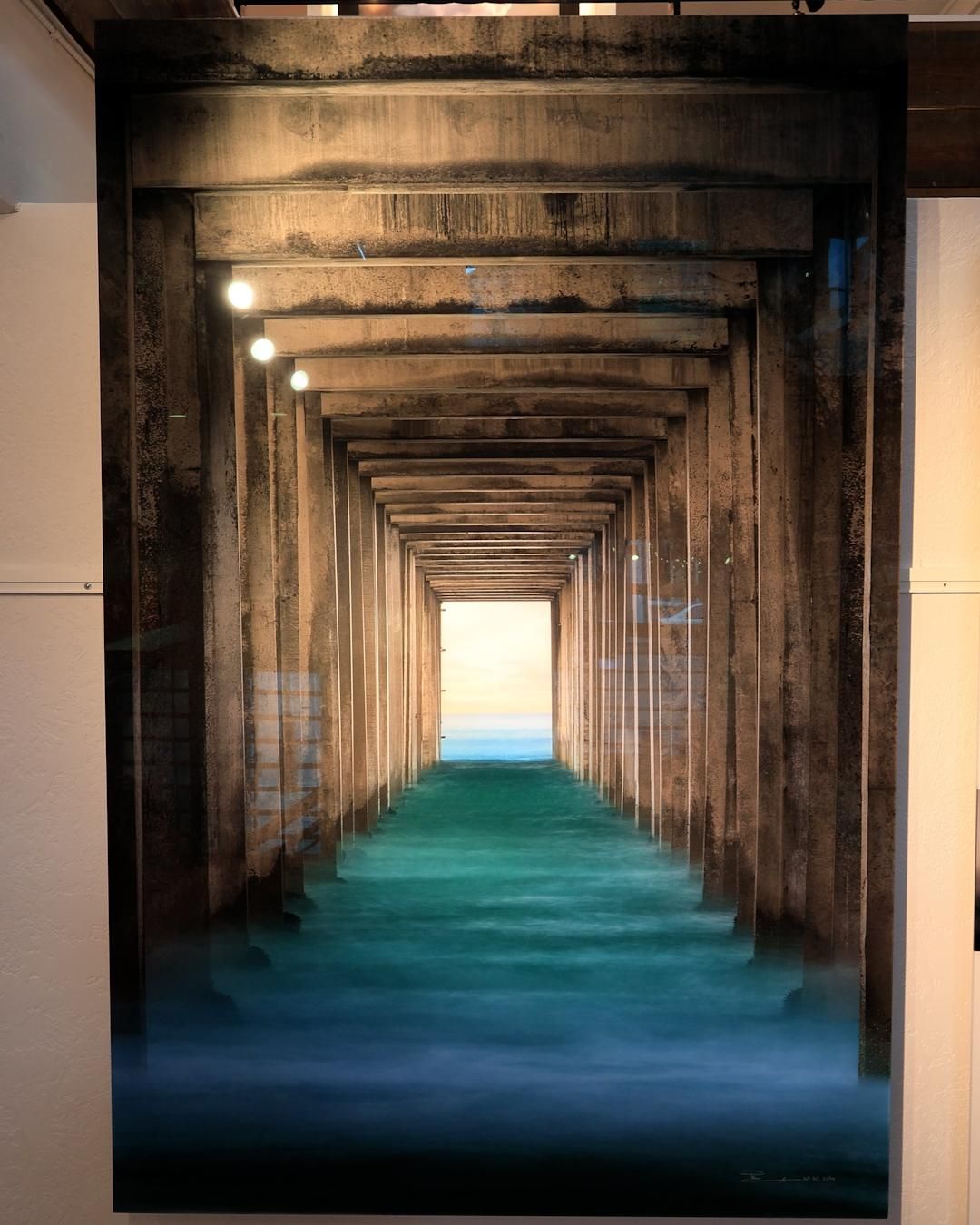 Peter Lik's 'Ocean Window' — vertical photograph looking down the tunnel of pilings beneath Scripps Pier in La Jolla, with misty turquoise water flowing toward a bright ocean horizon.