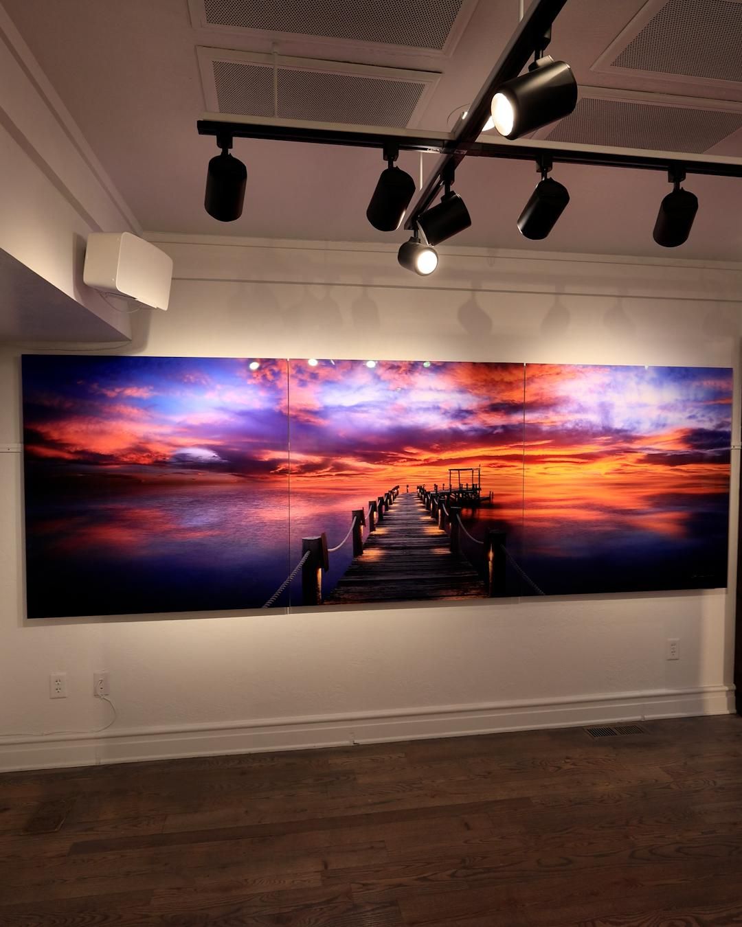 Peter Lik's 'Heavenly Night' displayed as a three-panel acrylic print — a wooden dock with rope railings stretching into calm water beneath a fiery orange, pink, and purple sunset.