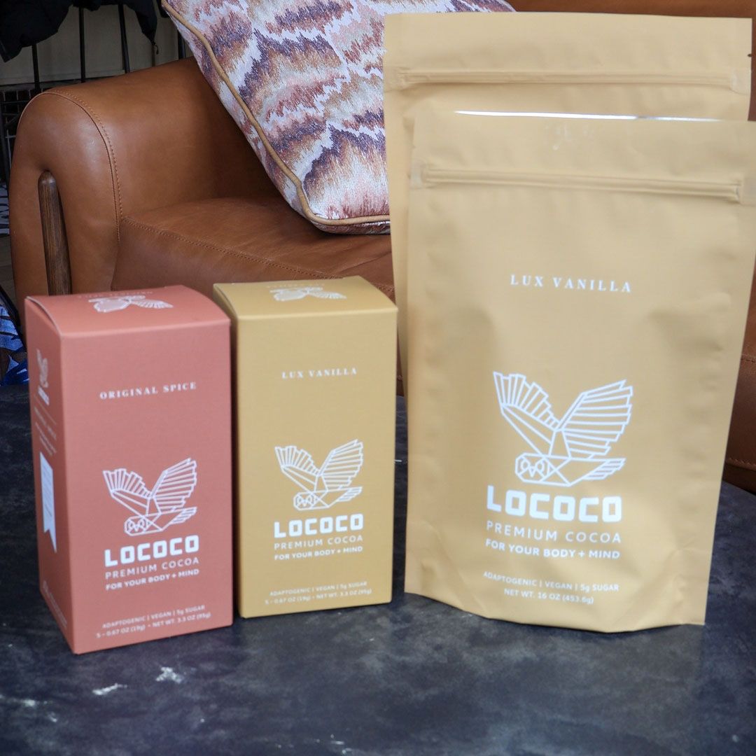 A close-up display of Lococo premium hot cocoa products sitting on a dark table, including an Original Spice box, a Lux Vanilla box, and a large Lux Vanilla pouch, all featuring the Lococo geometric owl logo.