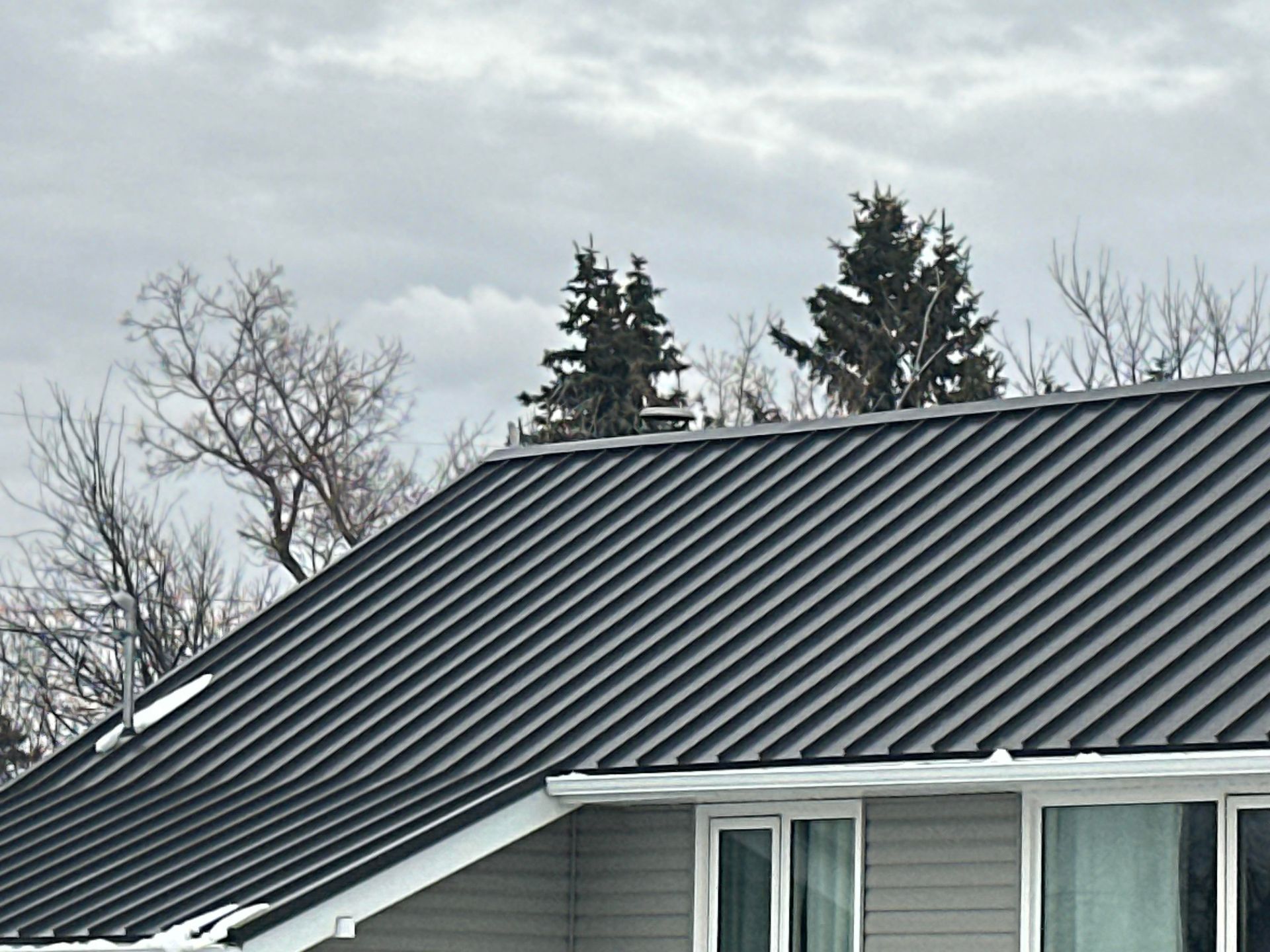 Dark metal roof on a house with white trim under a cloudy sky, with trees in the background