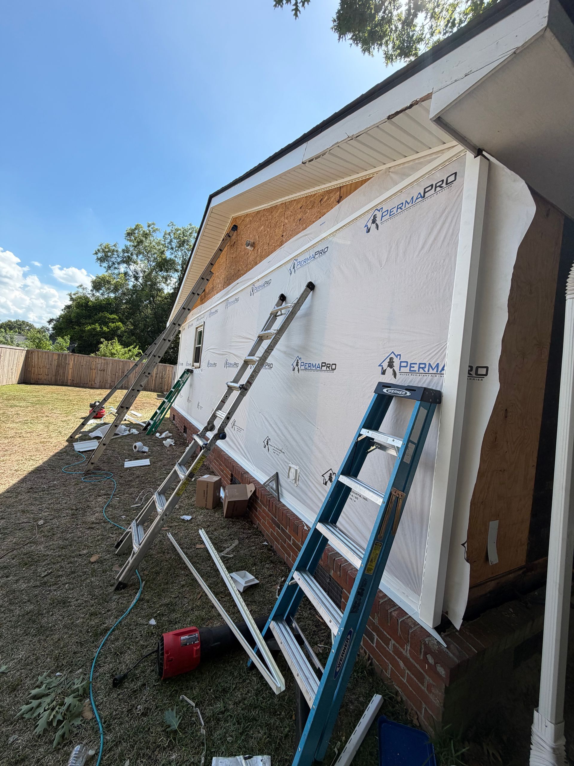 A Charleston Roofs and Windows inspection manager walking a Wando, SC homeowner through a line-item roof replacement estimate on a covered back porch, with a new Atlas shingle roof overhead and the pine tree line of the Cainhoy peninsula behind the yard. The image represents roof replacement cost in Wando SC, priced per square for asphalt shingles, metal and flat roofs, with free inspections and 0% financing for 12 months on approved credit.