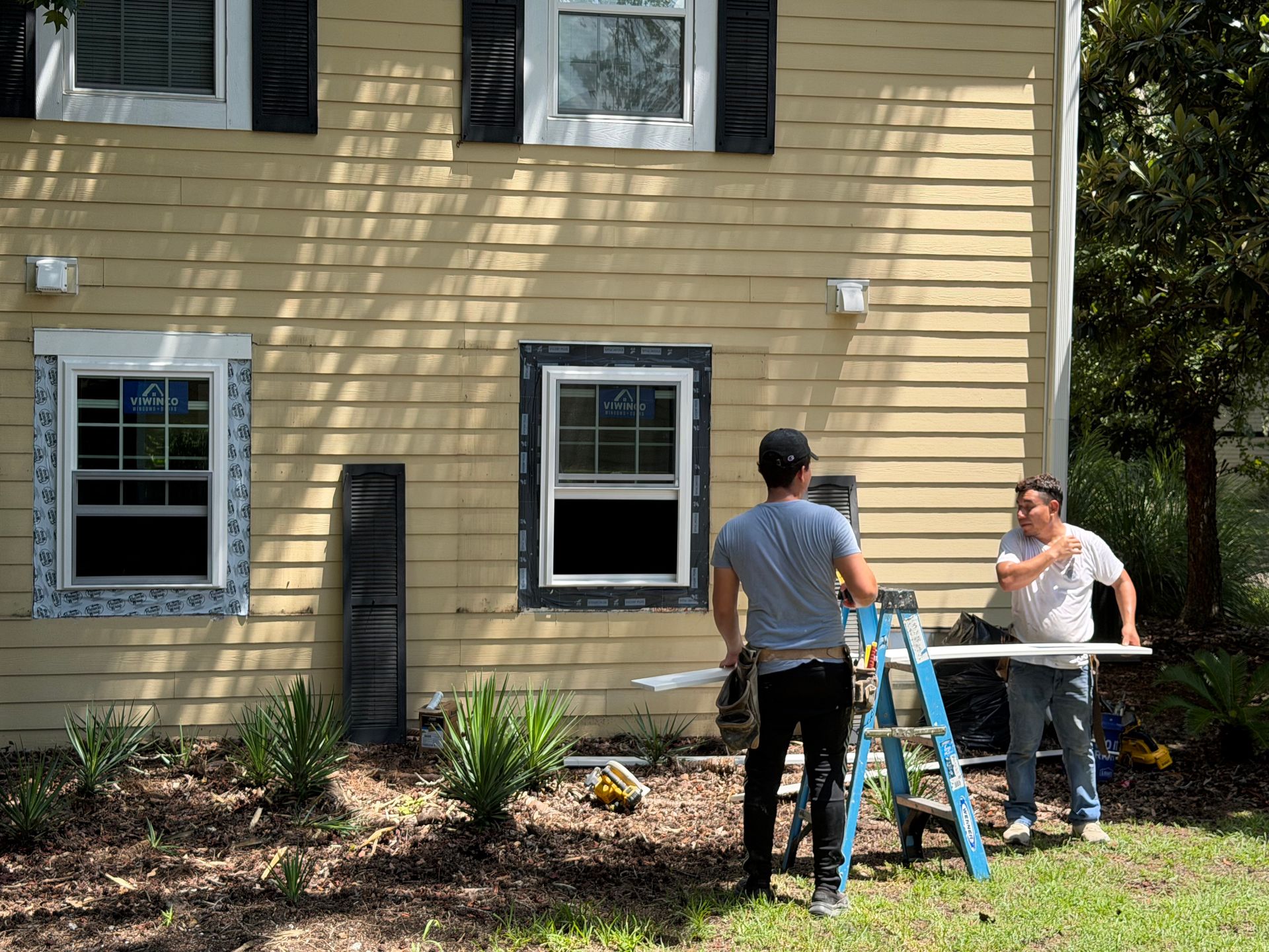 Two people stand by a house wall with a ladder, looking at the windows and siding.