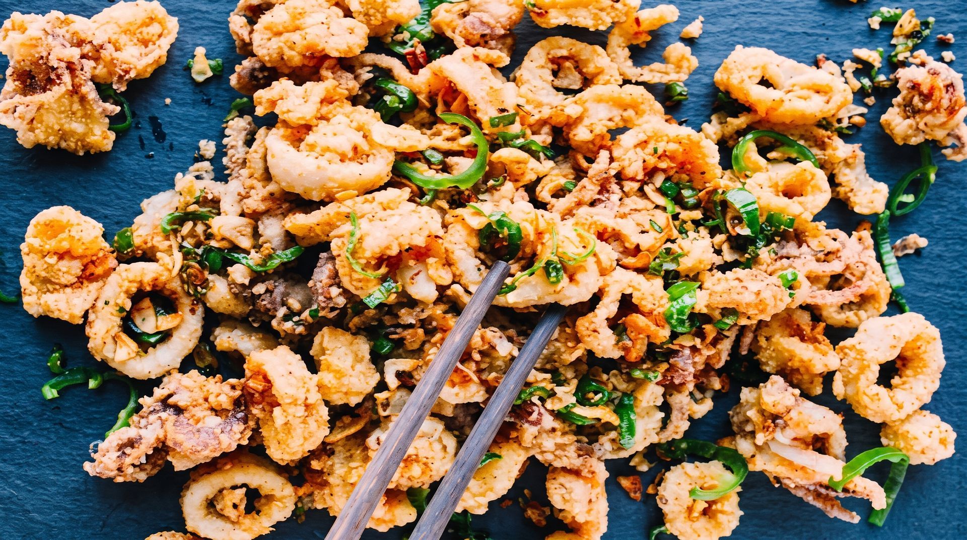Chopsticks stirring crispy fried calamari with herbs on a dark blue plate