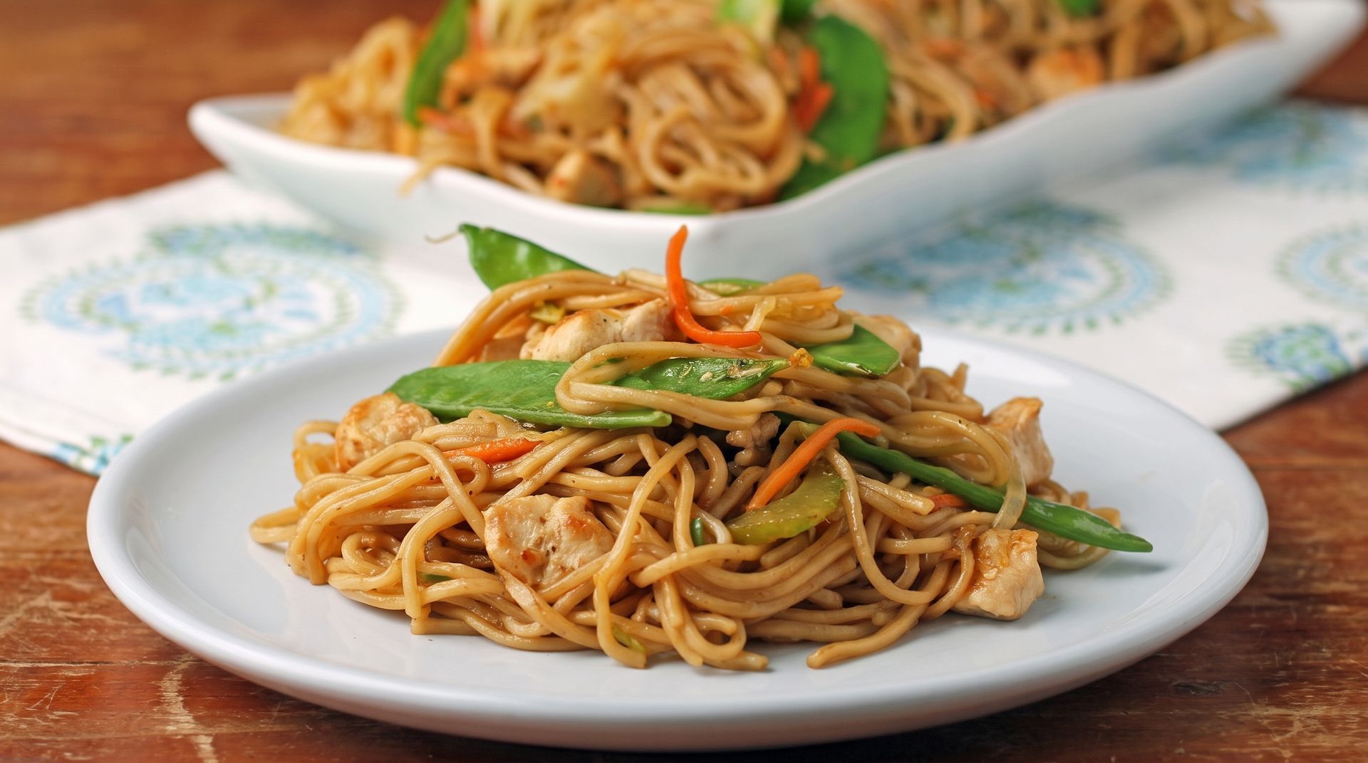 Two plates of stir-fried noodles topped with green vegetables on a patterned tablecloth