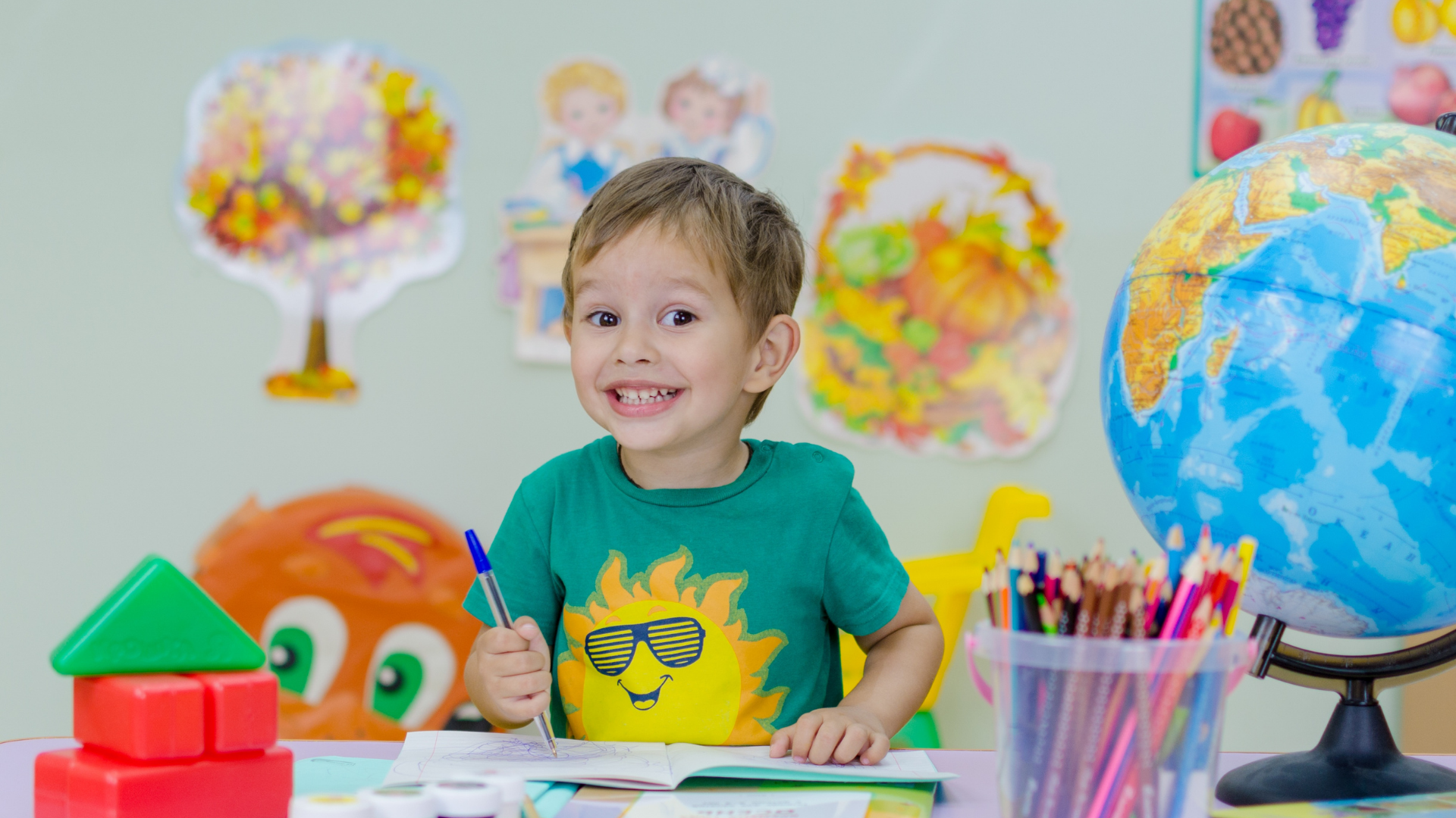 Smiling child in a green shirt at a desk, holding a pen in a classroom with colorful decorations.