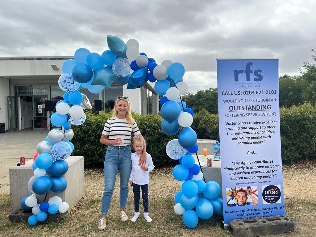 Woman and child pose under blue balloon arch at an outdoor event.