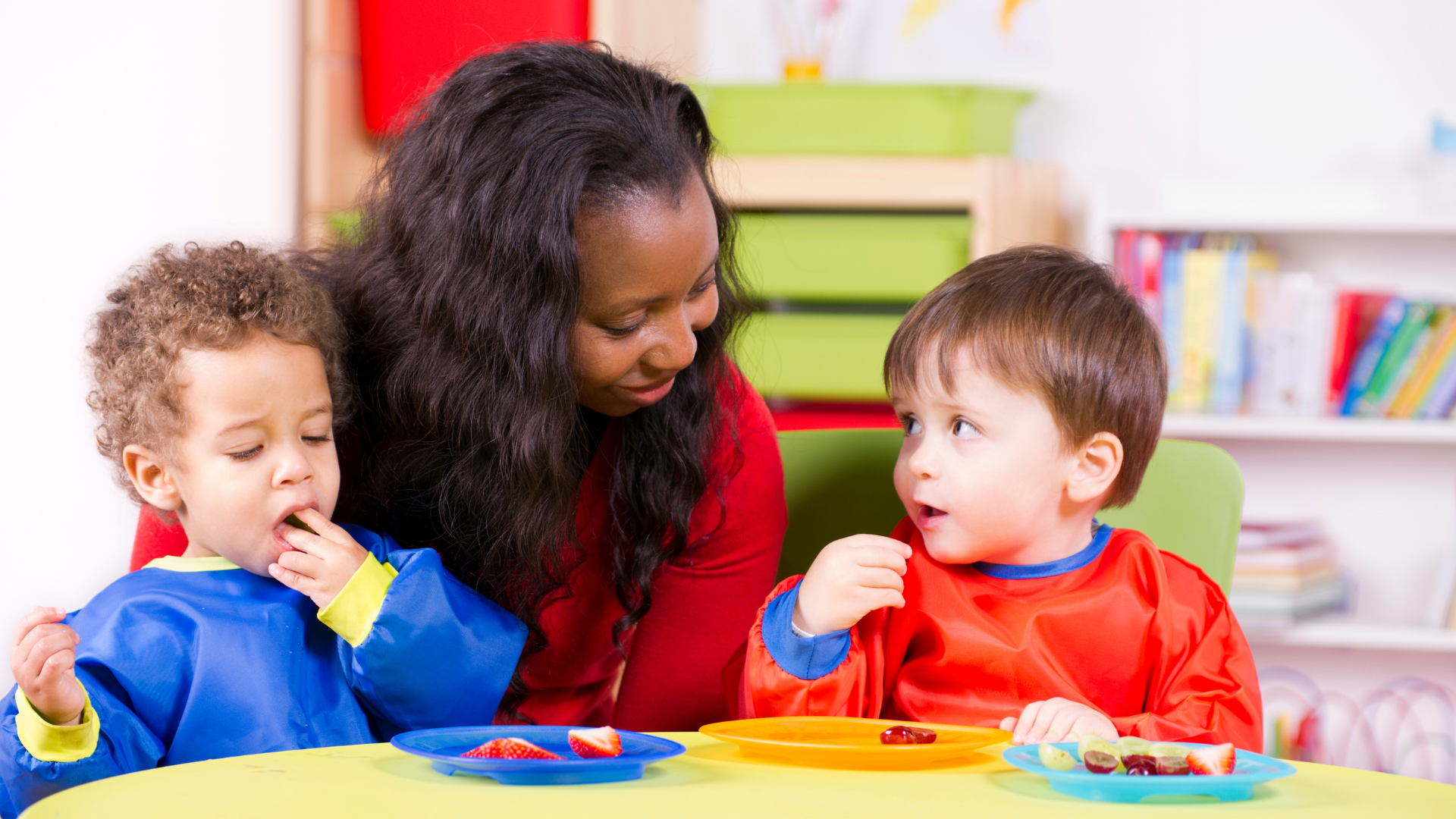 Woman watching two children eating at a table. Children wear bibs.