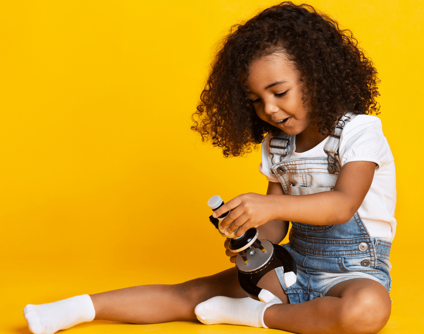 Young Black girl, smiles while playing with a microscope, yellow backdrop.
