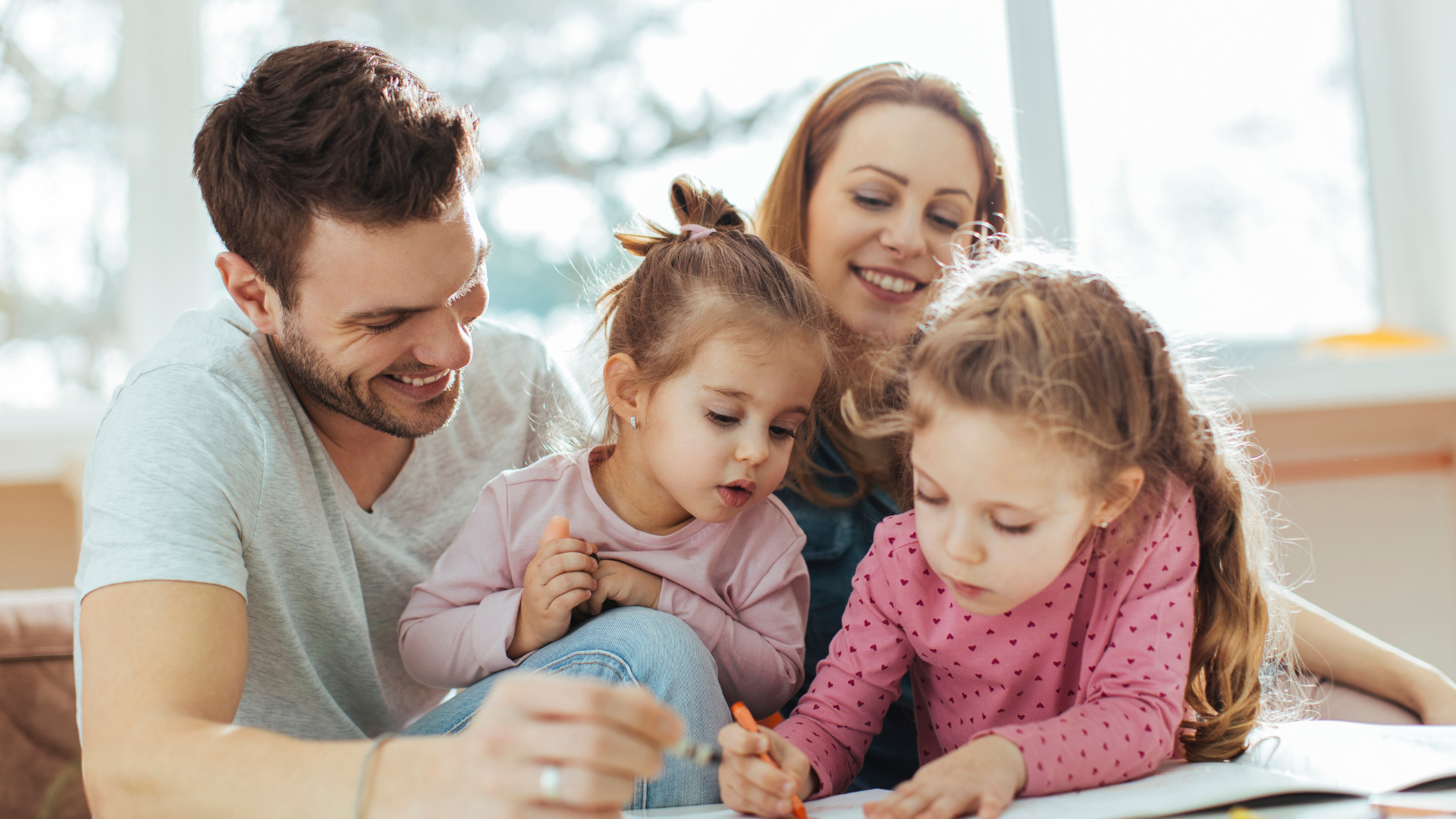 Family of four drawing together, smiling. Light-filled room, dad holding crayon, two girls, mom watching.