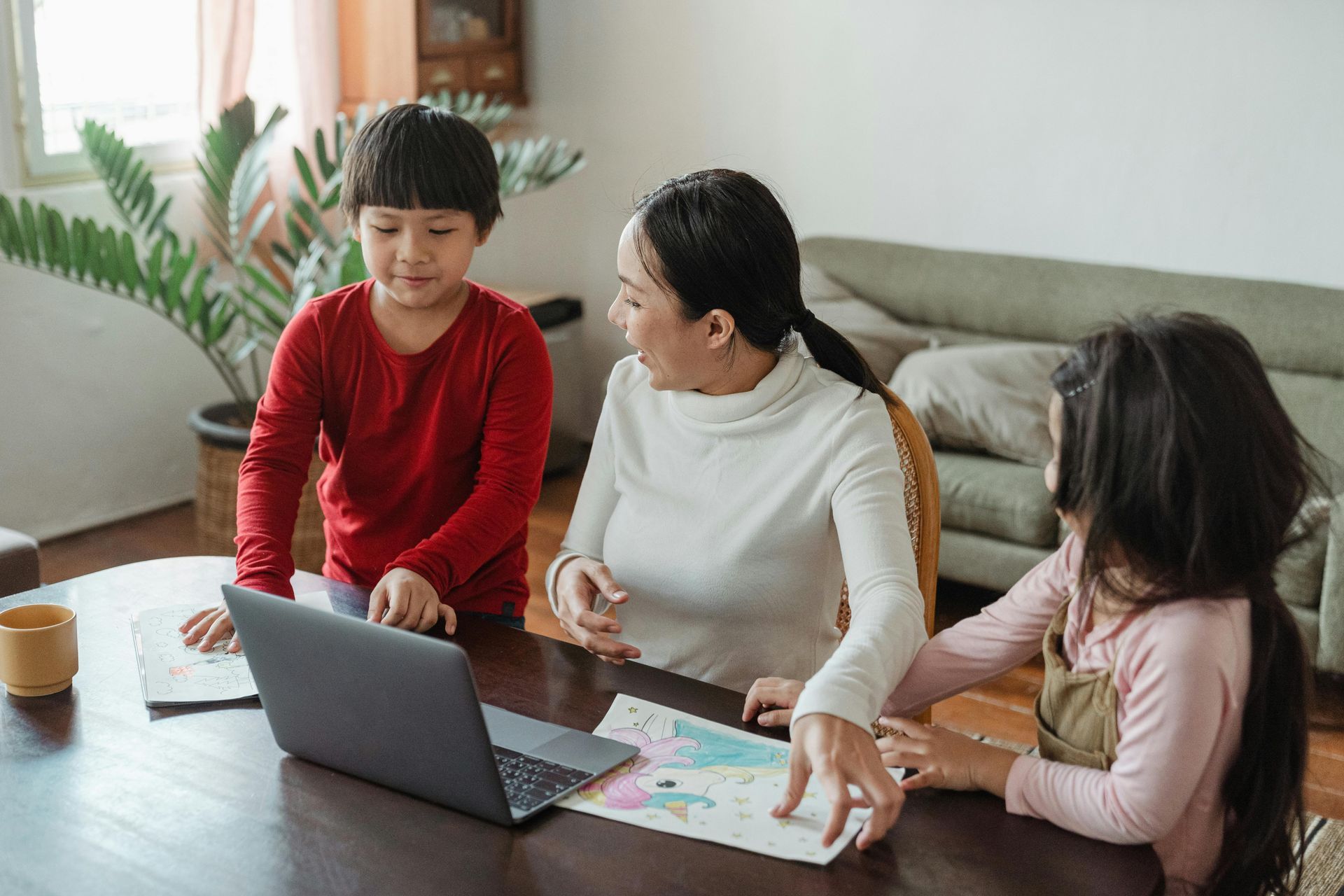 Woman, two children at a table, looking at a laptop. They're working on coloring pages.