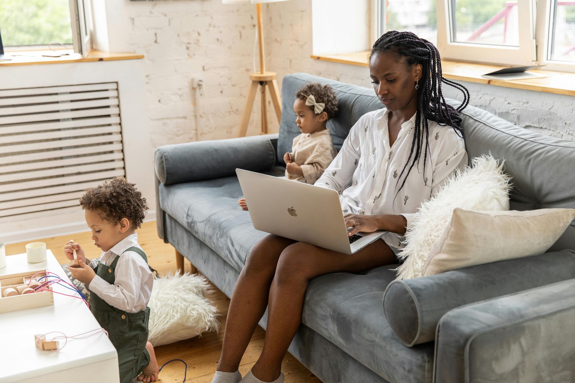 Woman working on laptop on couch with two children in a living room.
